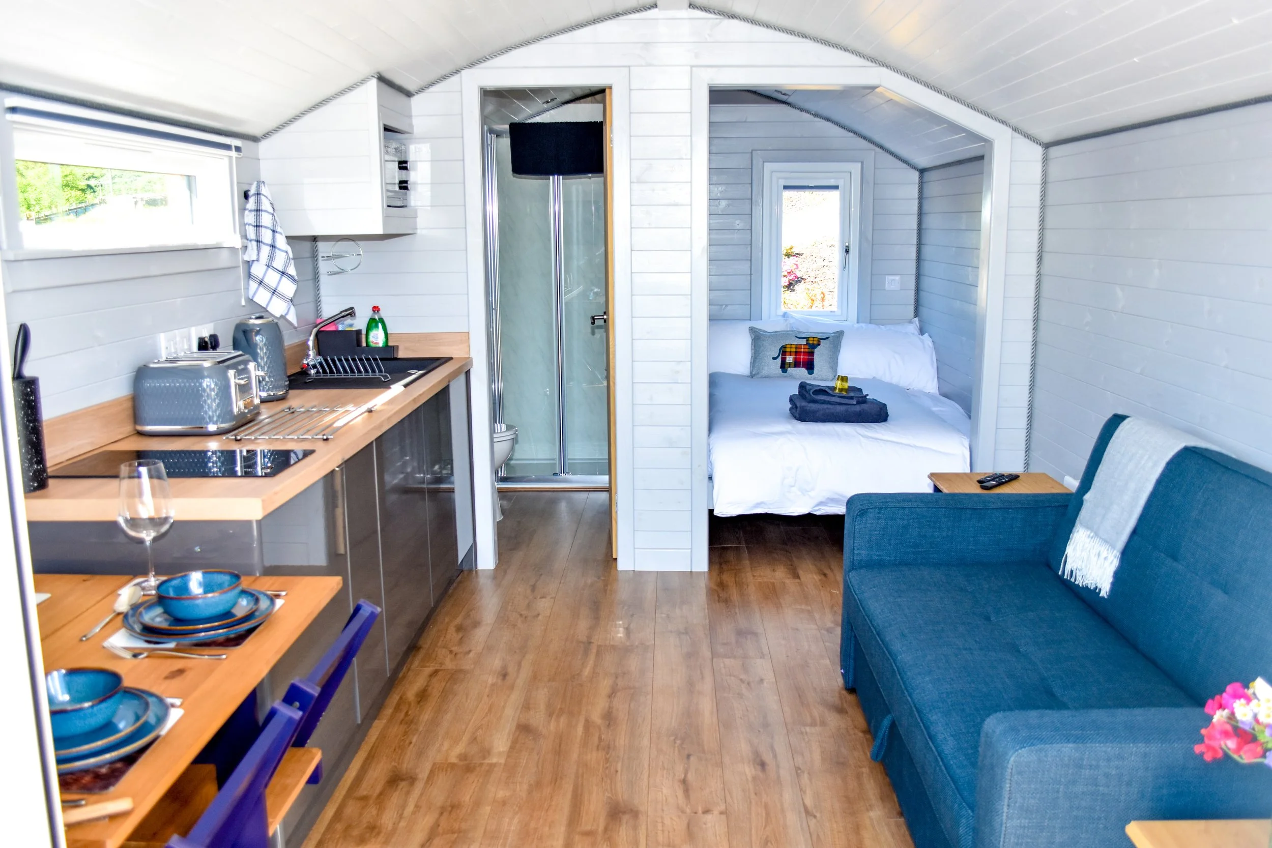 Interior of a compact modern tiny home with a kitchen area, small bedroom, and a sitting area with a blue sofa.