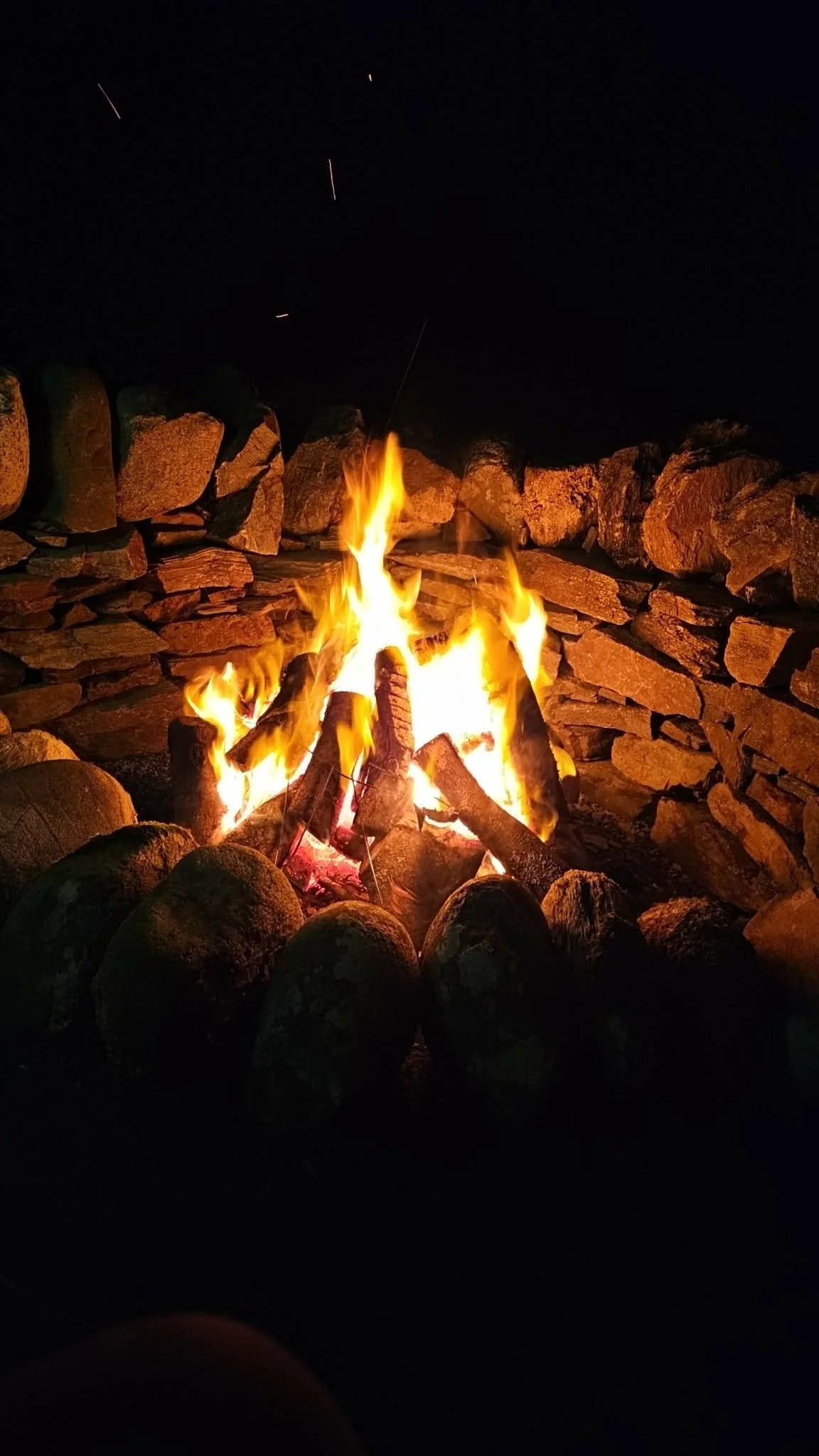 A campfire surrounded by rocks at night, with flames and sparks rising into the dark sky.