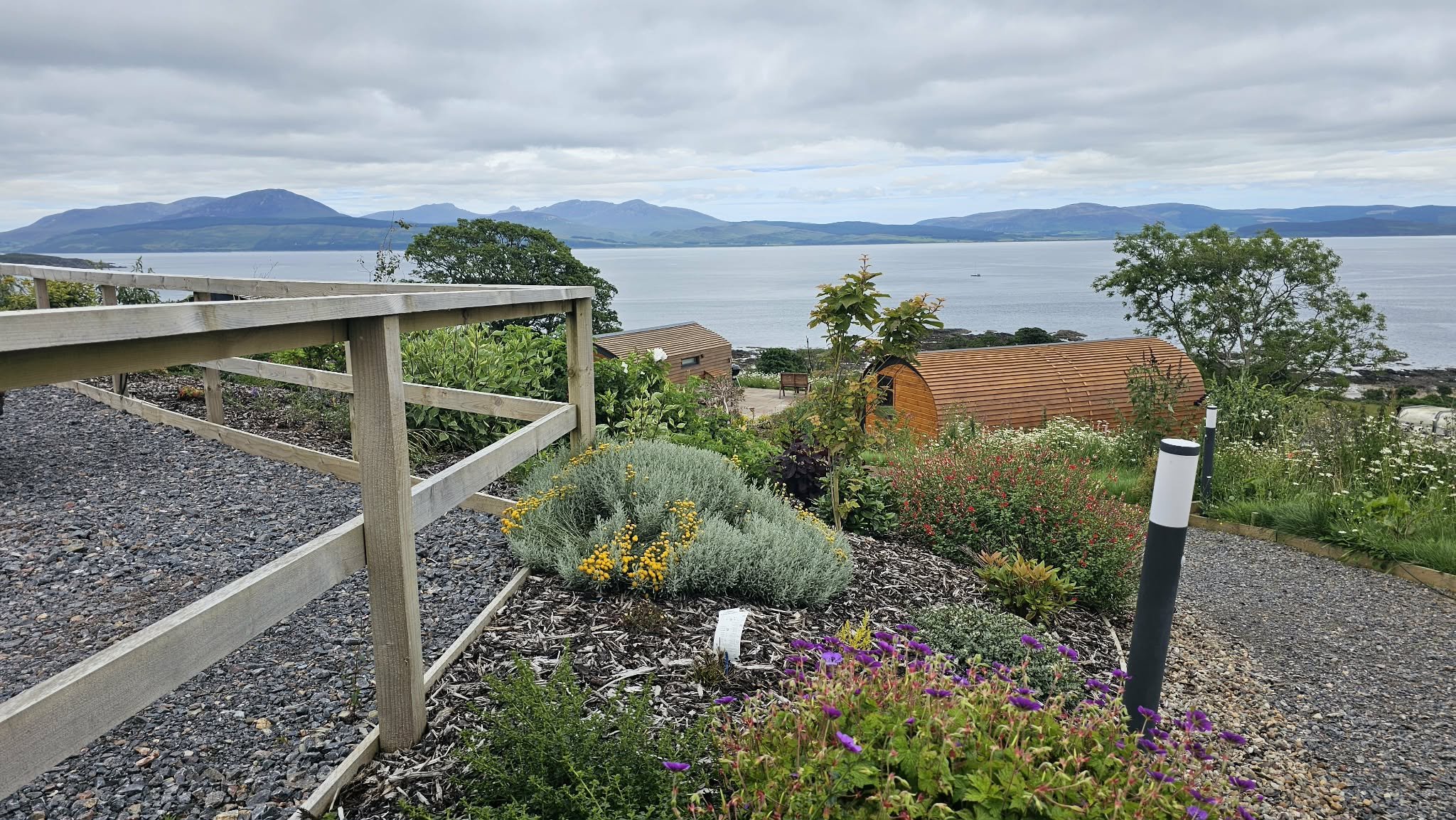 Scenic view of a garden with colorful flowers, a wooden shed, and a railing overlooking a body of water with mountains in the background, under a cloudy sky.