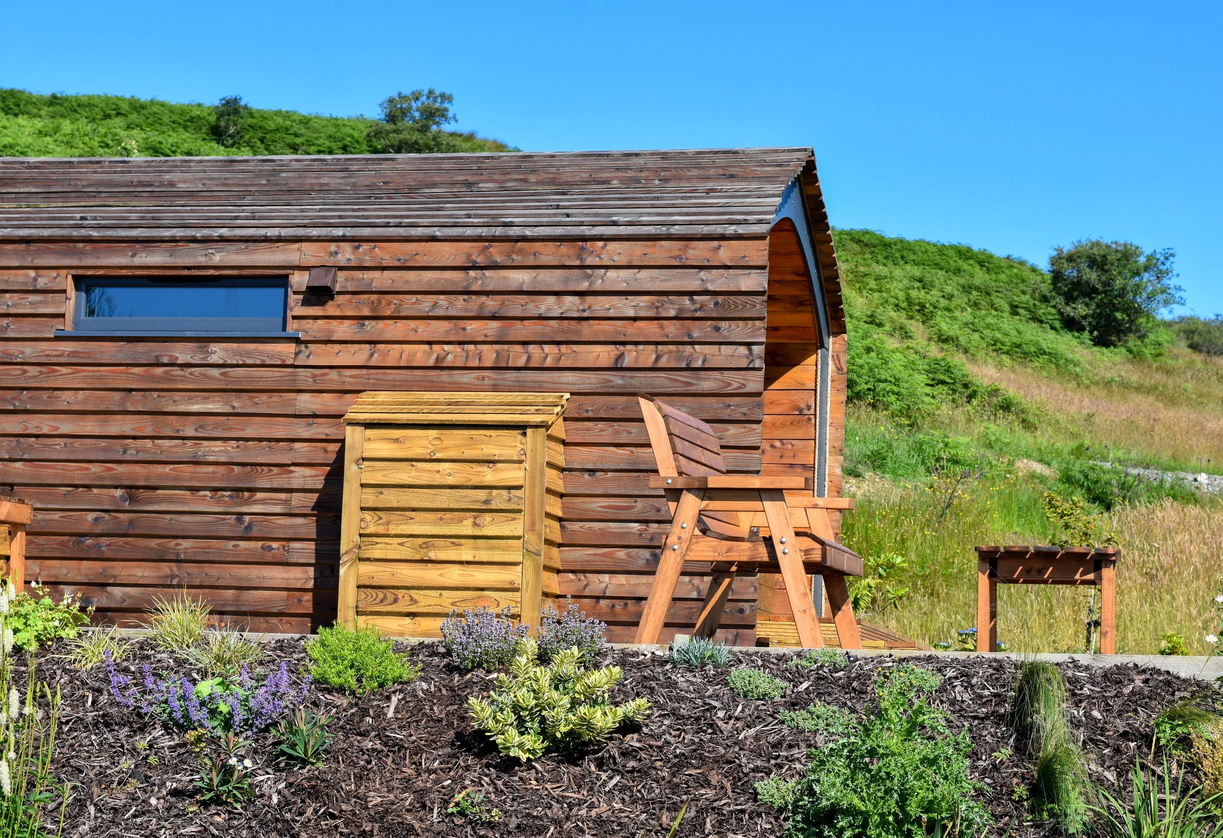 A wooden shed with a sloped roof and a small window, situated on a hillside with green vegetation, surrounded by a garden with various plants and flowers.