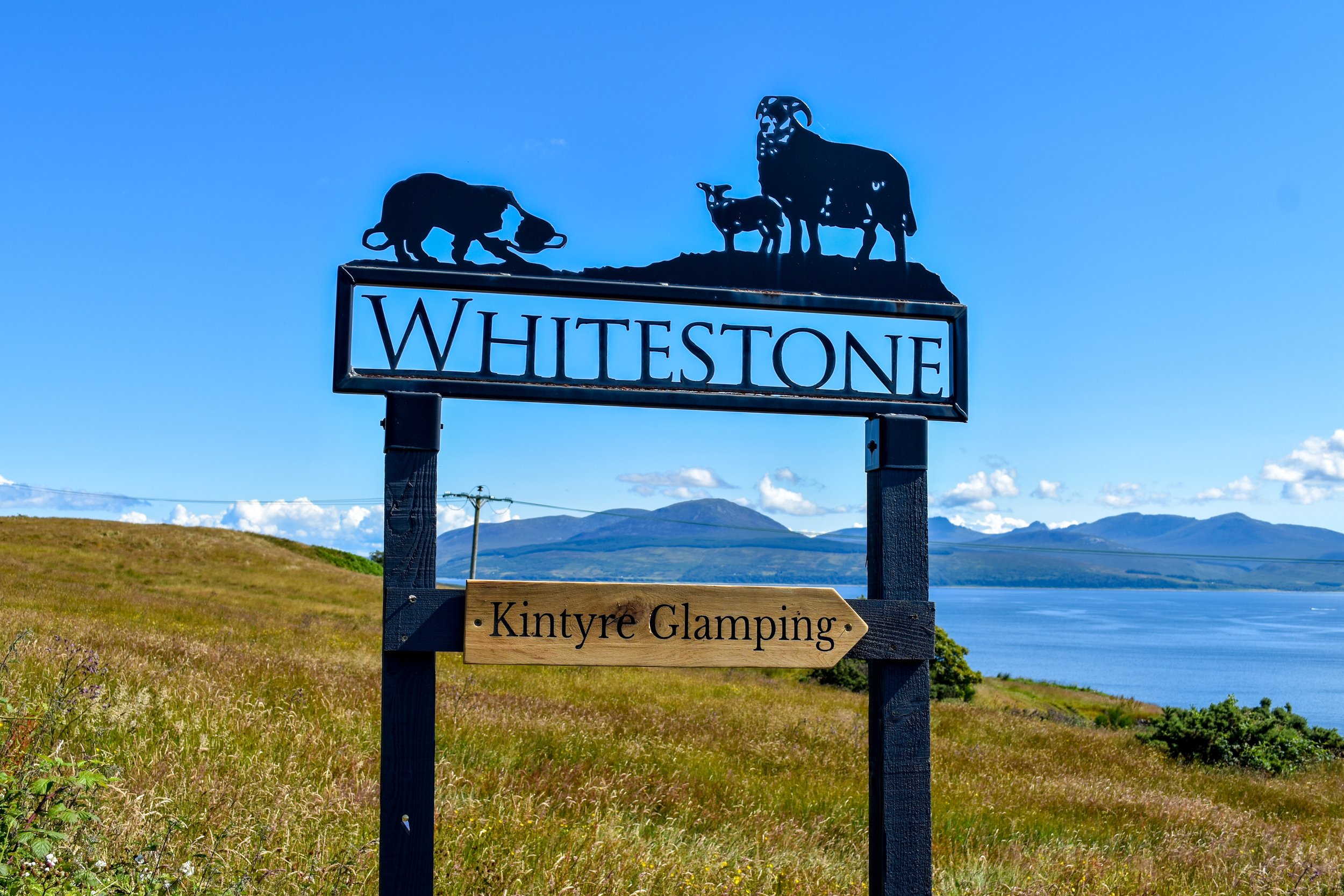 Sign for Whitestone with silhouettes of animals on top and a sign for Kintyre Glamping, overlooking a landscape with grass, water, and mountains in the background.