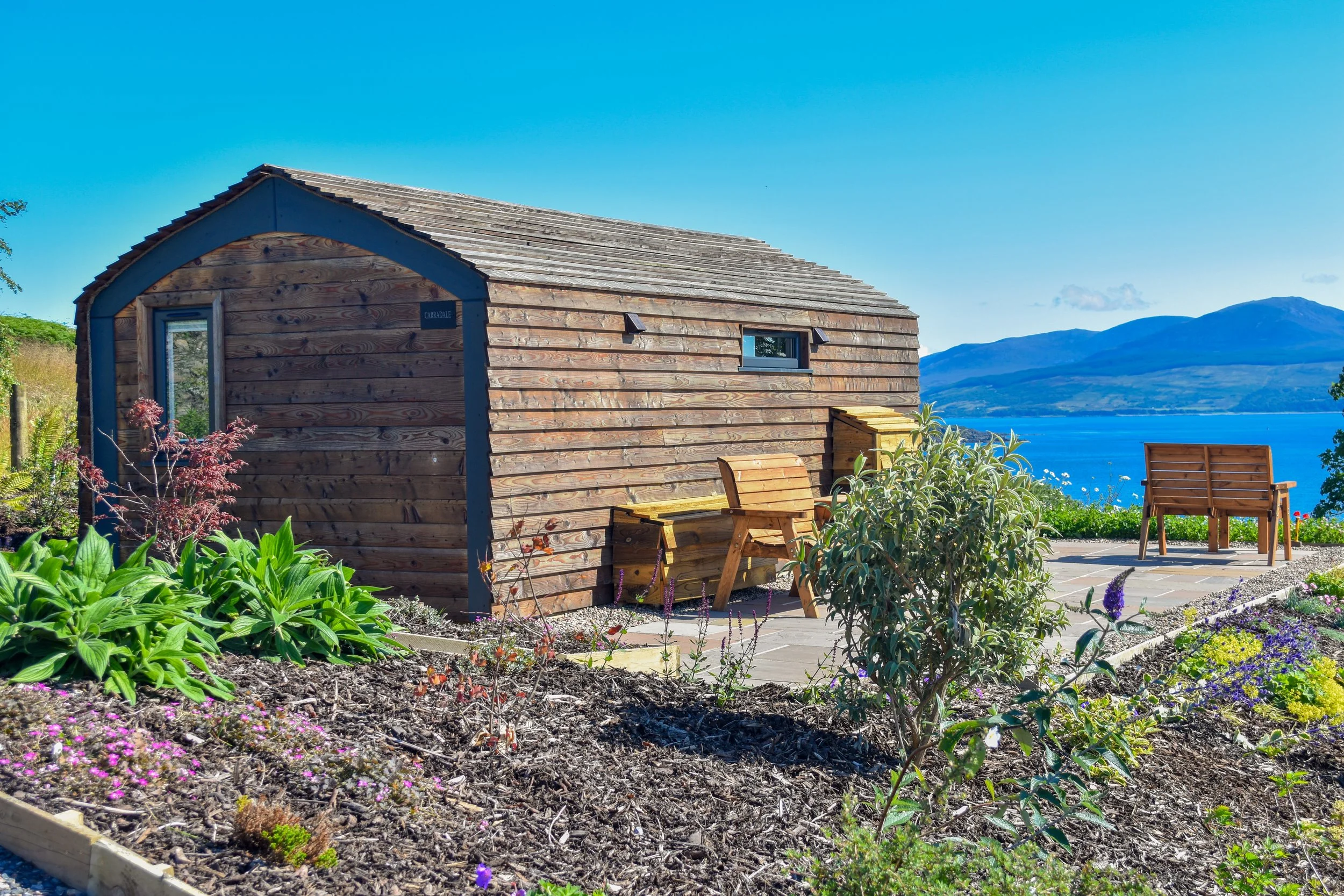 A wooden cabin on a hill with colorful flowers and plants in the garden, overlooking a blue lake and mountains in the background under a clear sky.