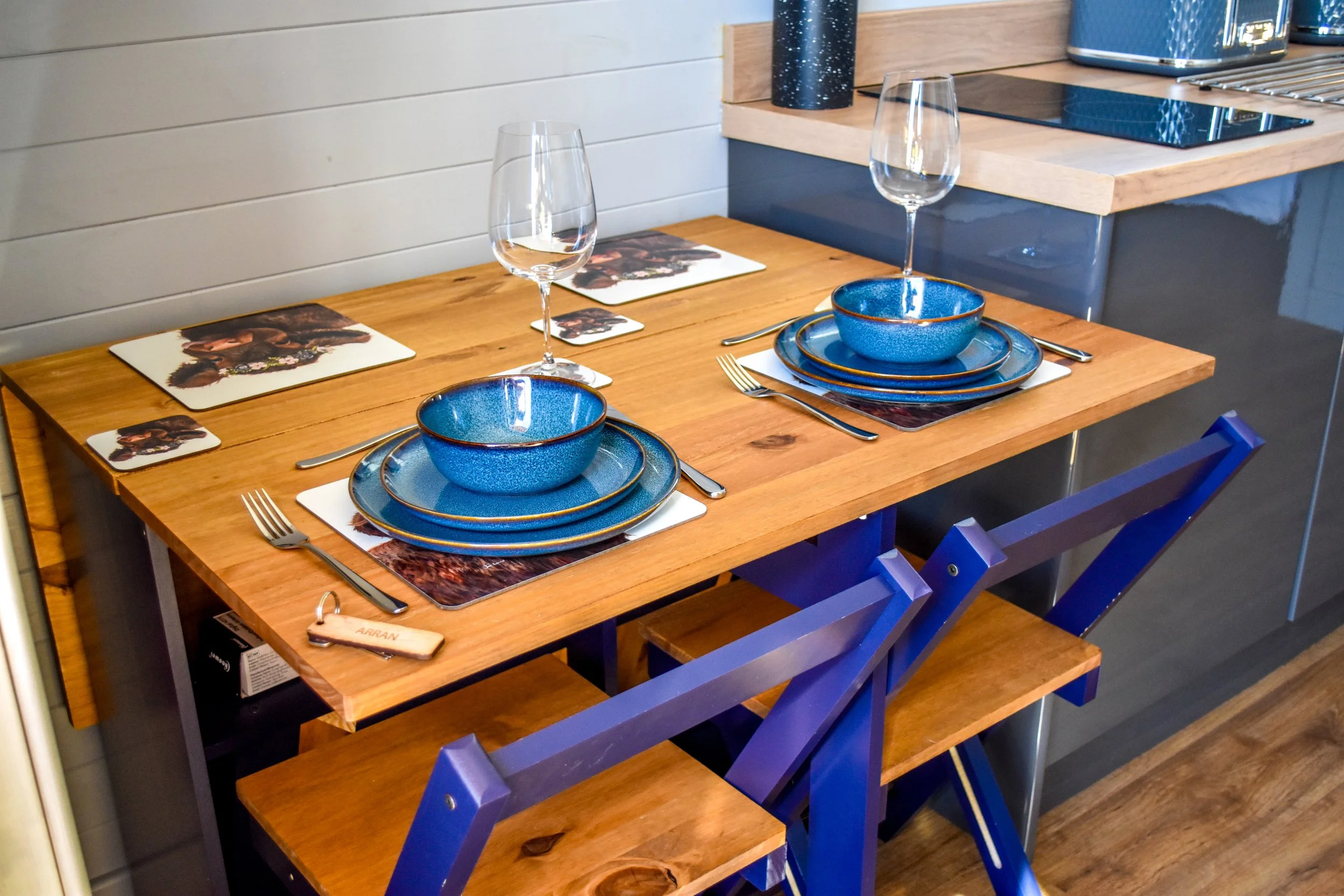 A dining table set for two with blue dinnerware, wine glasses, silverware, and printed placemats, situated in a modern kitchen or dining area.