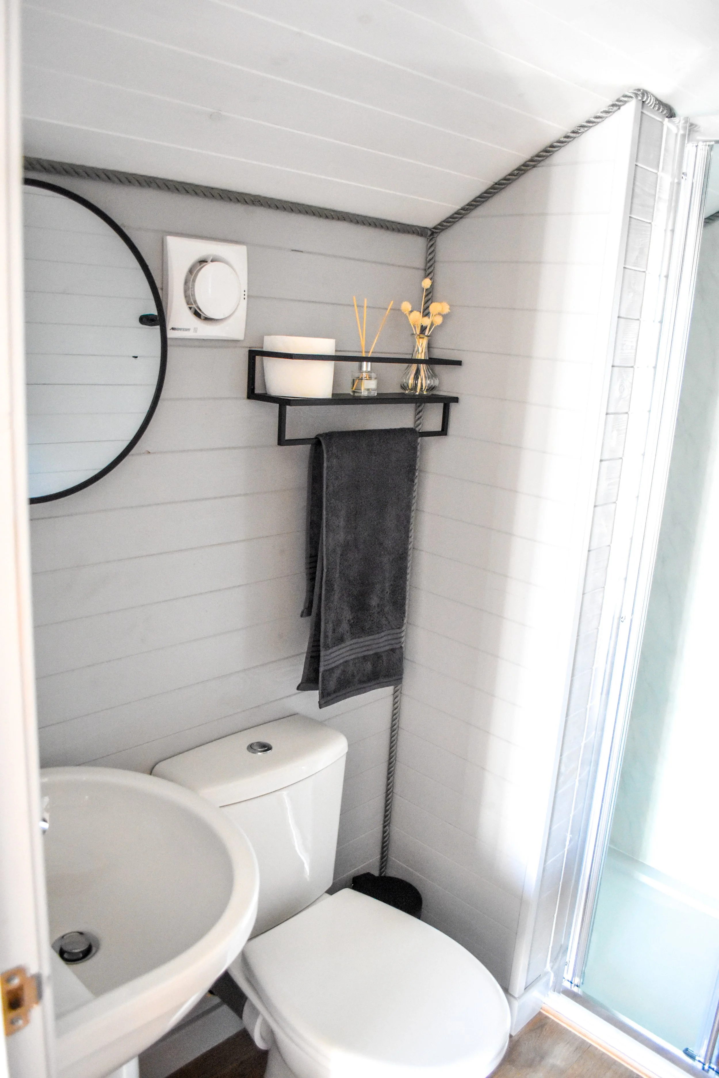 Small bathroom with white wall panels, a white toilet, a small white sink, a round mirror, a black shelf with a candle and decorative items, dark gray towel, and a glass shower enclosure.