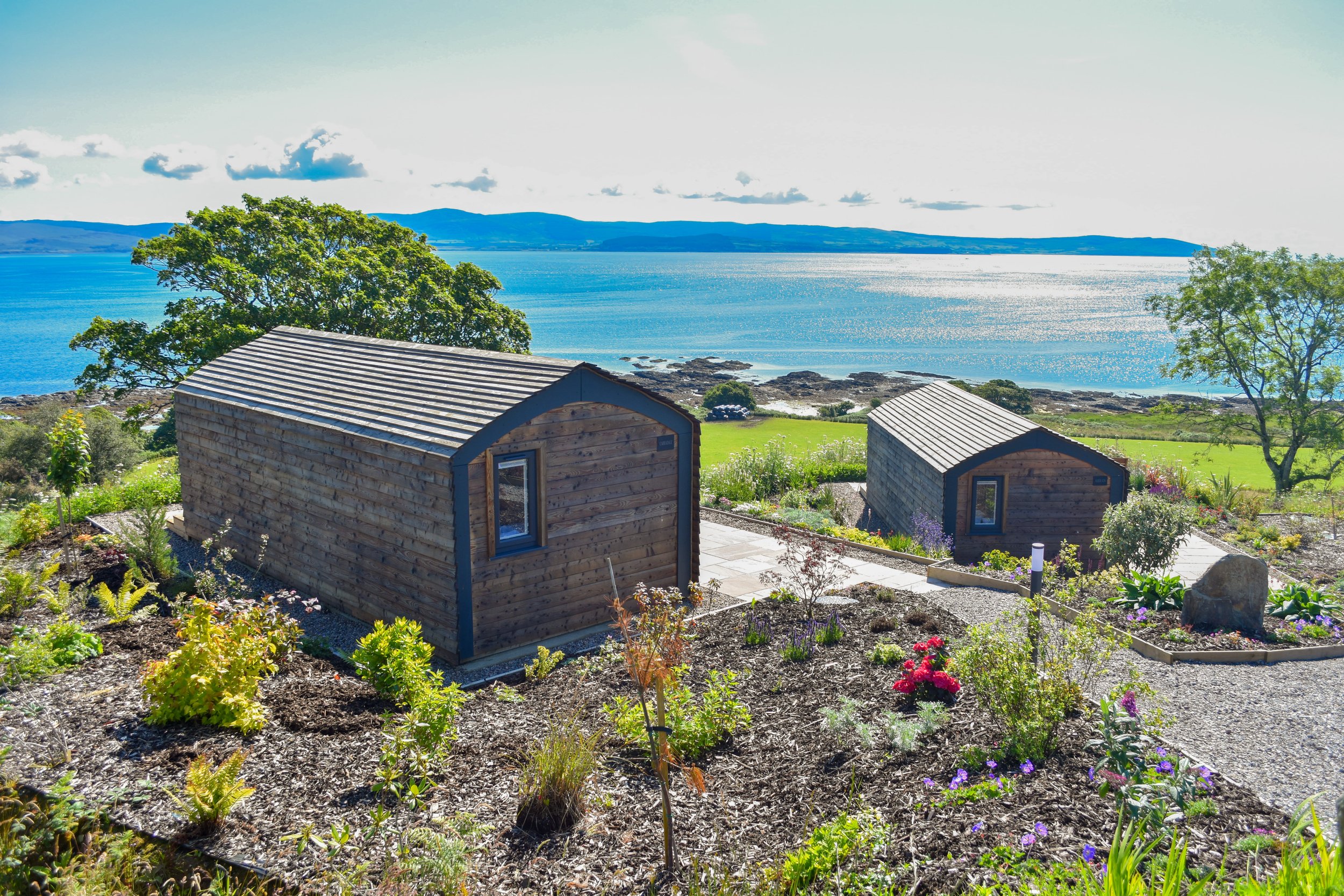 View of two wooden cabins on a hill with a garden, overlooking a lake and distant mountains under a partly cloudy sky.