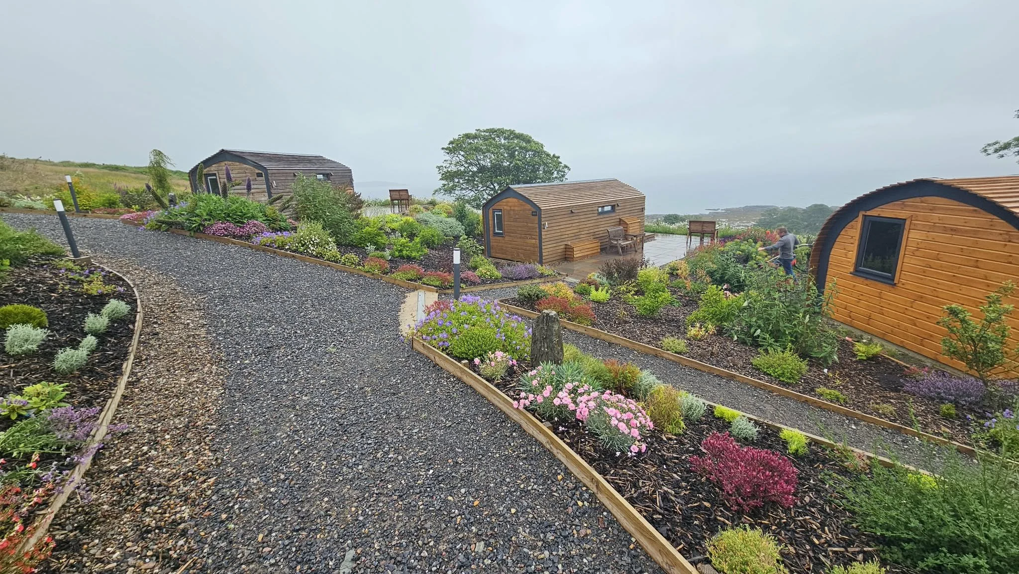 A gravel pathway winding through colorful flower beds and small wooden cabins with a scenic view in the background.