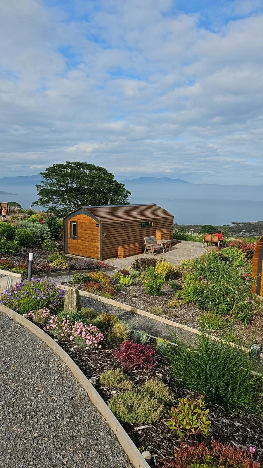 A scenic view of a small wooden cabin on a hilltop with a garden, overlooking a body of water with mountains in the distance and a partly cloudy sky.