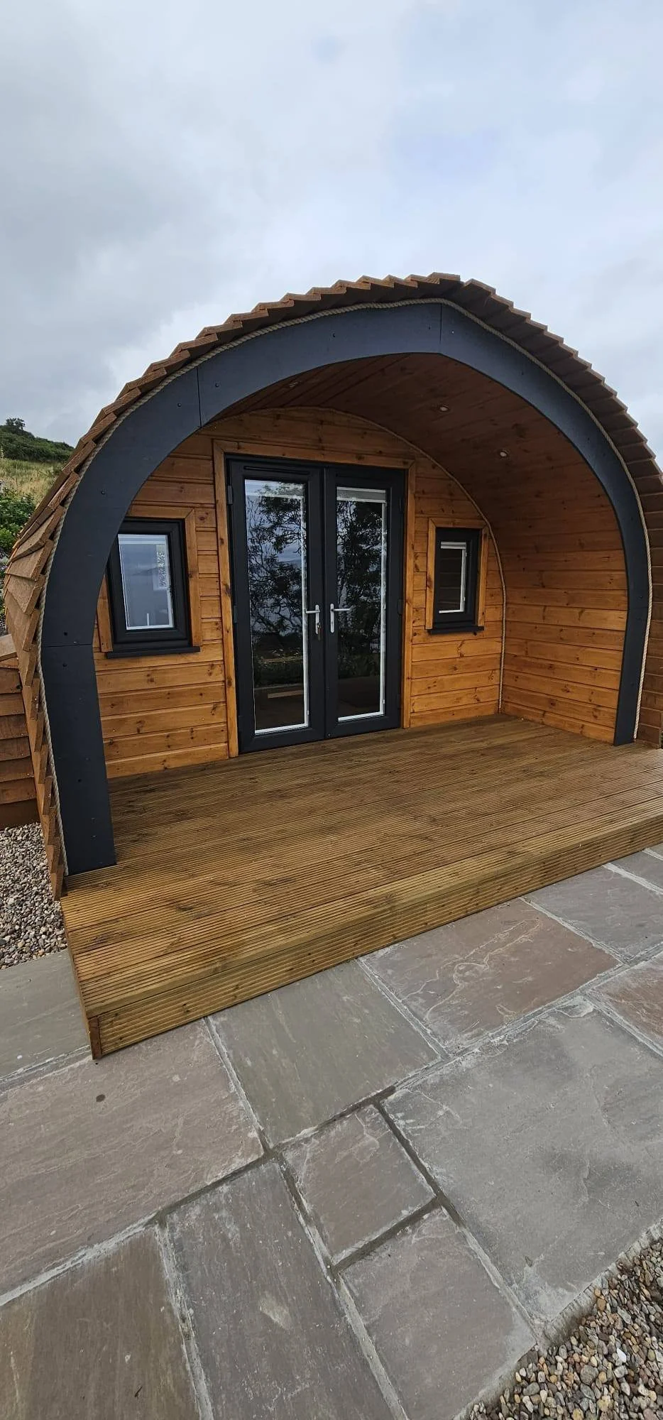 Small wooden house with a curved roof, double glass doors, and two small windows, set on a wooden deck, under an overcast sky.