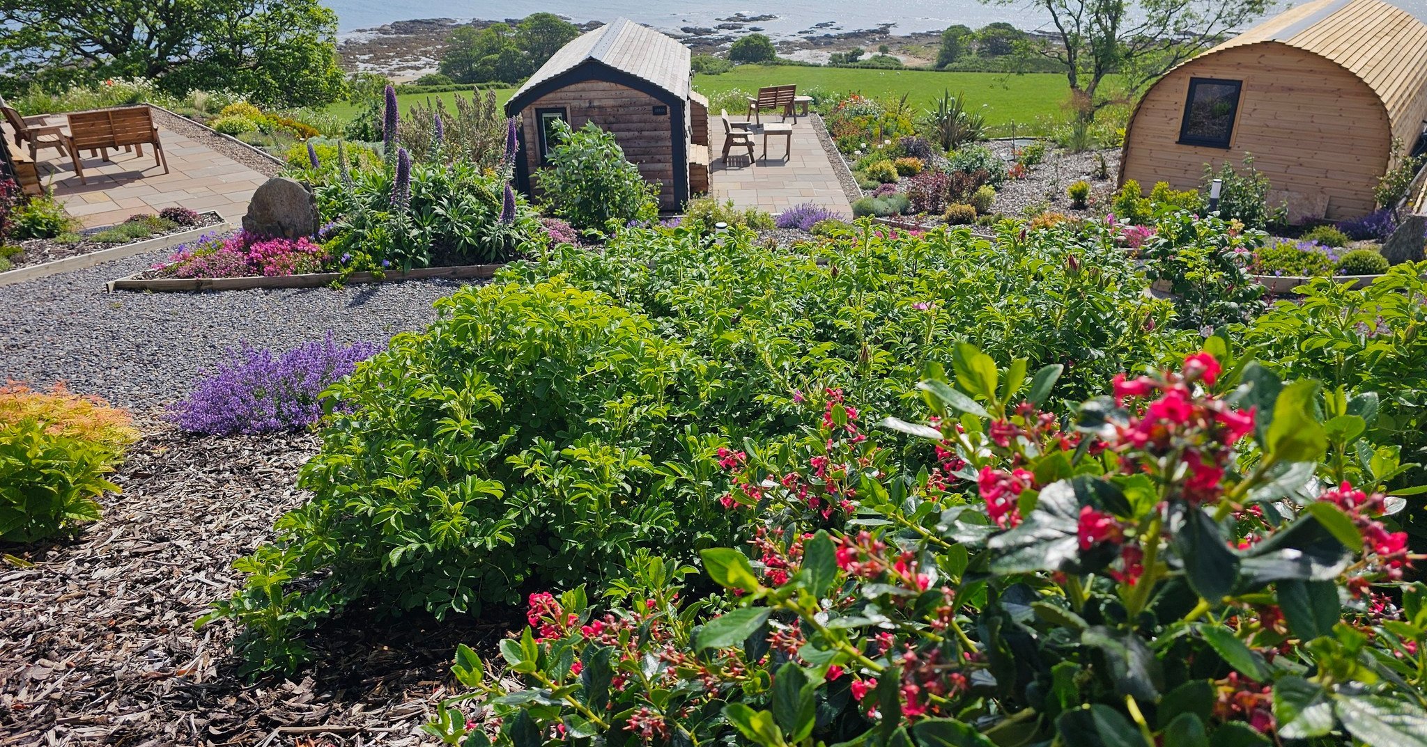 A vibrant garden with lush green plants, colorful flowers, and two wooden structures, set against a scenic landscape with rolling hills and a cloudy sky.