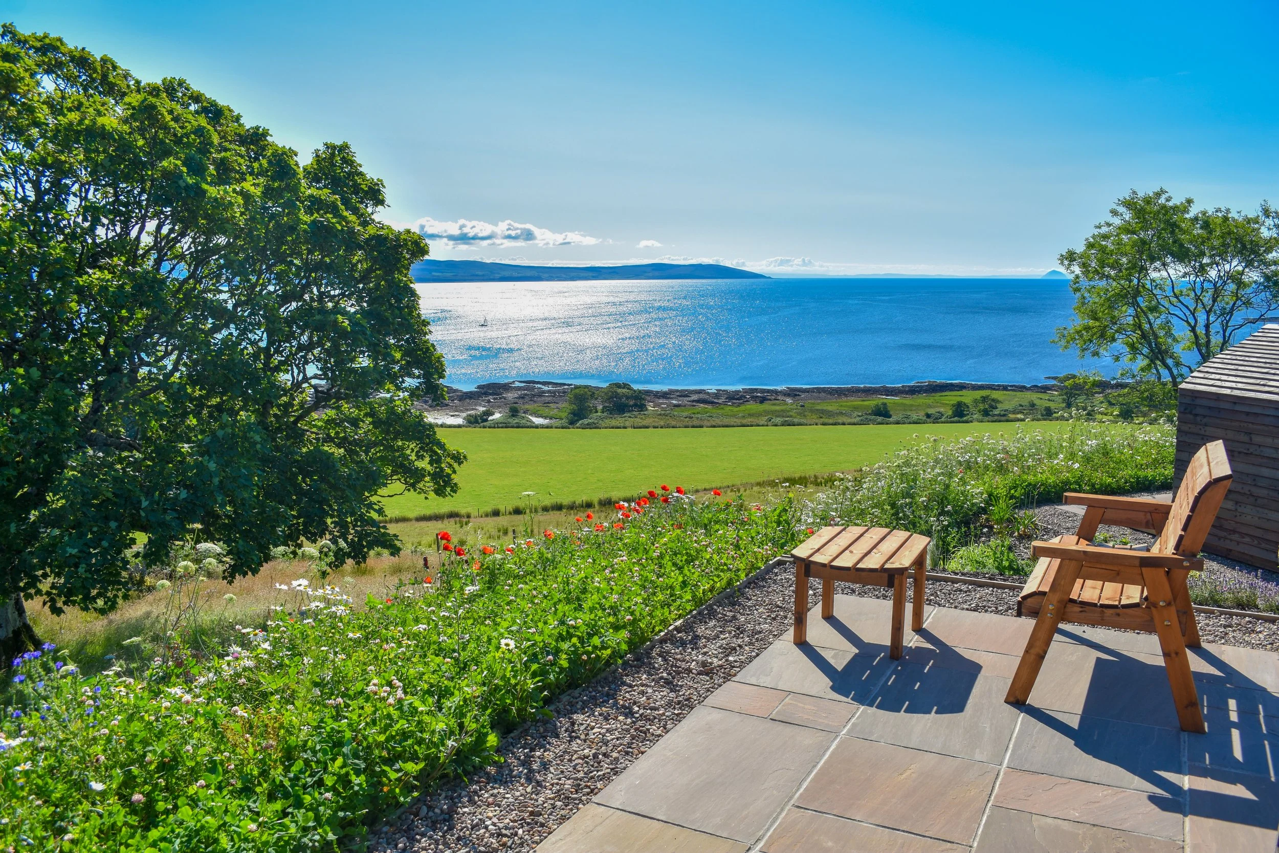 A scenic view from a patio with wooden chairs and a small table, overlooking a green field, colorful flowers, and a large body of water under a bright blue sky.