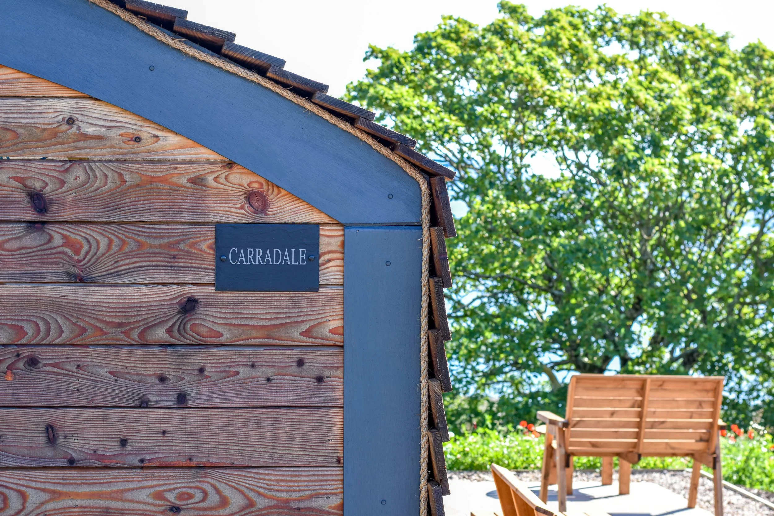 Close-up of a wooden shed with a black nameplate that says 'CARRADALE', with a large green tree and a wooden bench in the background, under a clear blue sky.