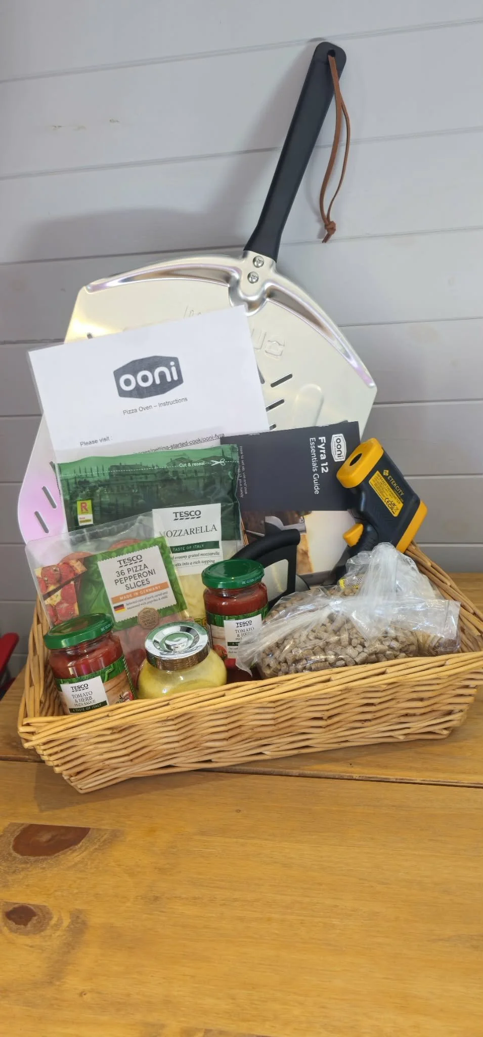 A wicker basket on a wooden table filled with pizza ingredients and tools, including jars of tomato sauce, shredded cheese, a bag of cereal, a pizza cutter, and an oven thermometer, with a pizza peel leaning against the wall behind.