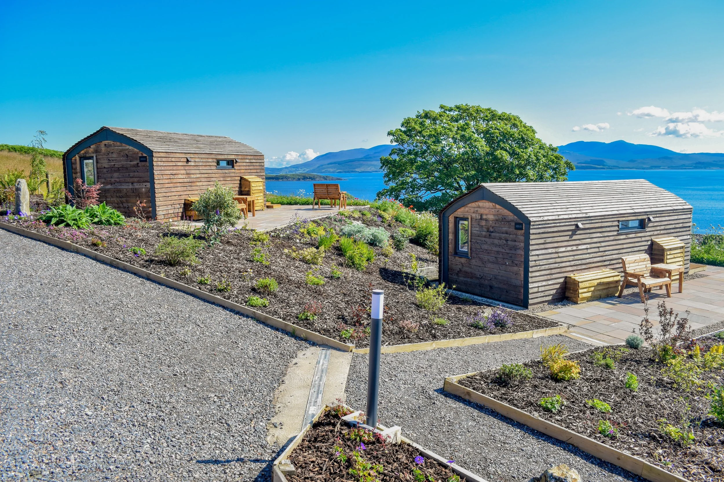 Two small wooden cabins on a hill with a view of a lake and mountains in the background, surrounded by a landscaped garden with flowers and plants.