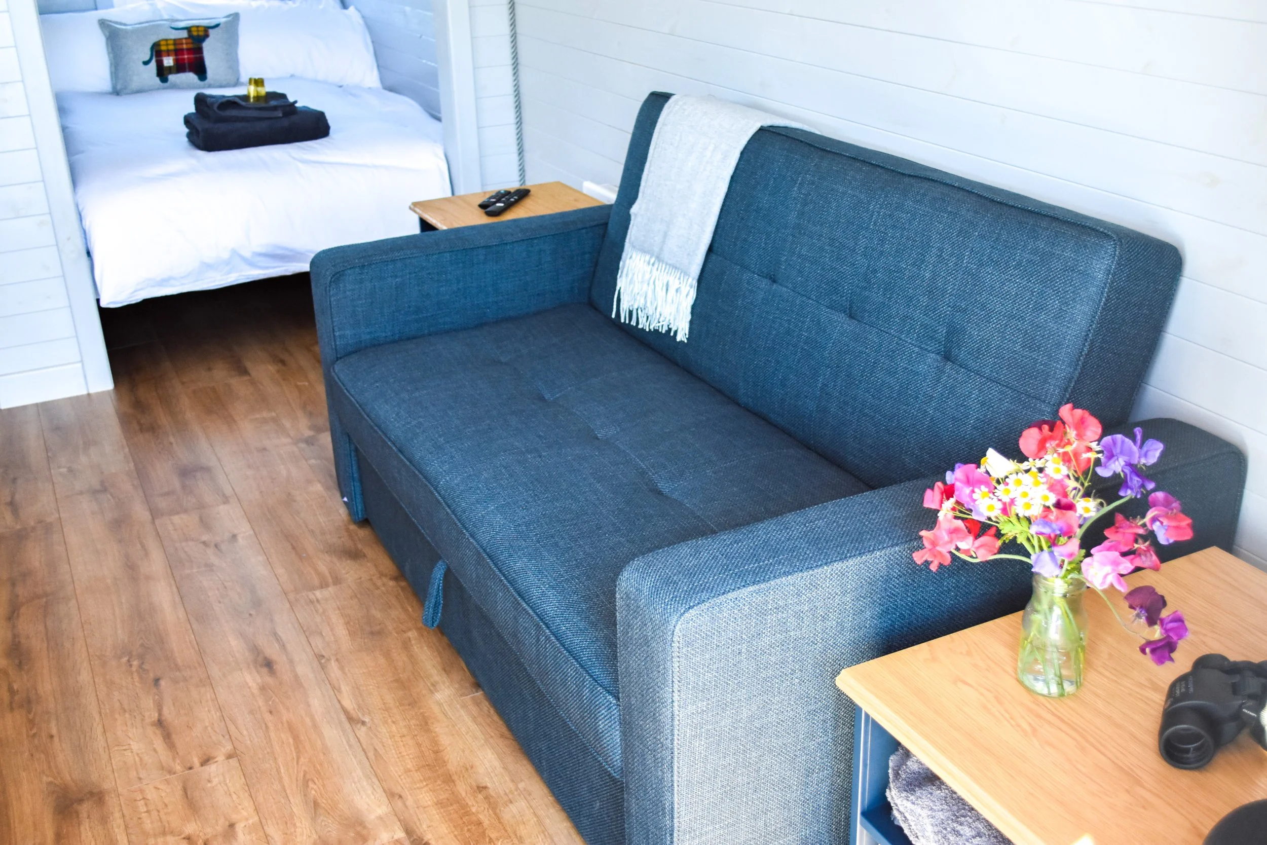 A cozy room with a blue fabric sofa, a wooden side table with a vase of pink and purple flowers, a bed with white linens and a decorative pillow, and a pair of binoculars on a wooden desk. The room has white wooden walls and wooden flooring.