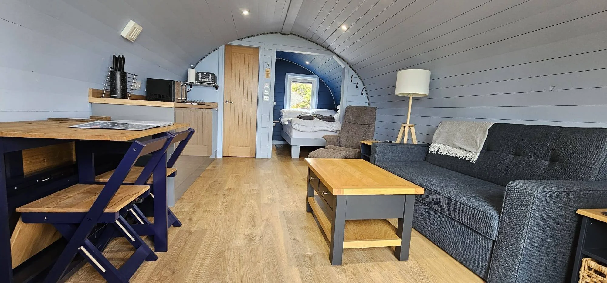 Interior of a compact living space with a small bedroom, kitchen area, and sitting area, featuring wooden flooring, light blue walls, and modern furniture.