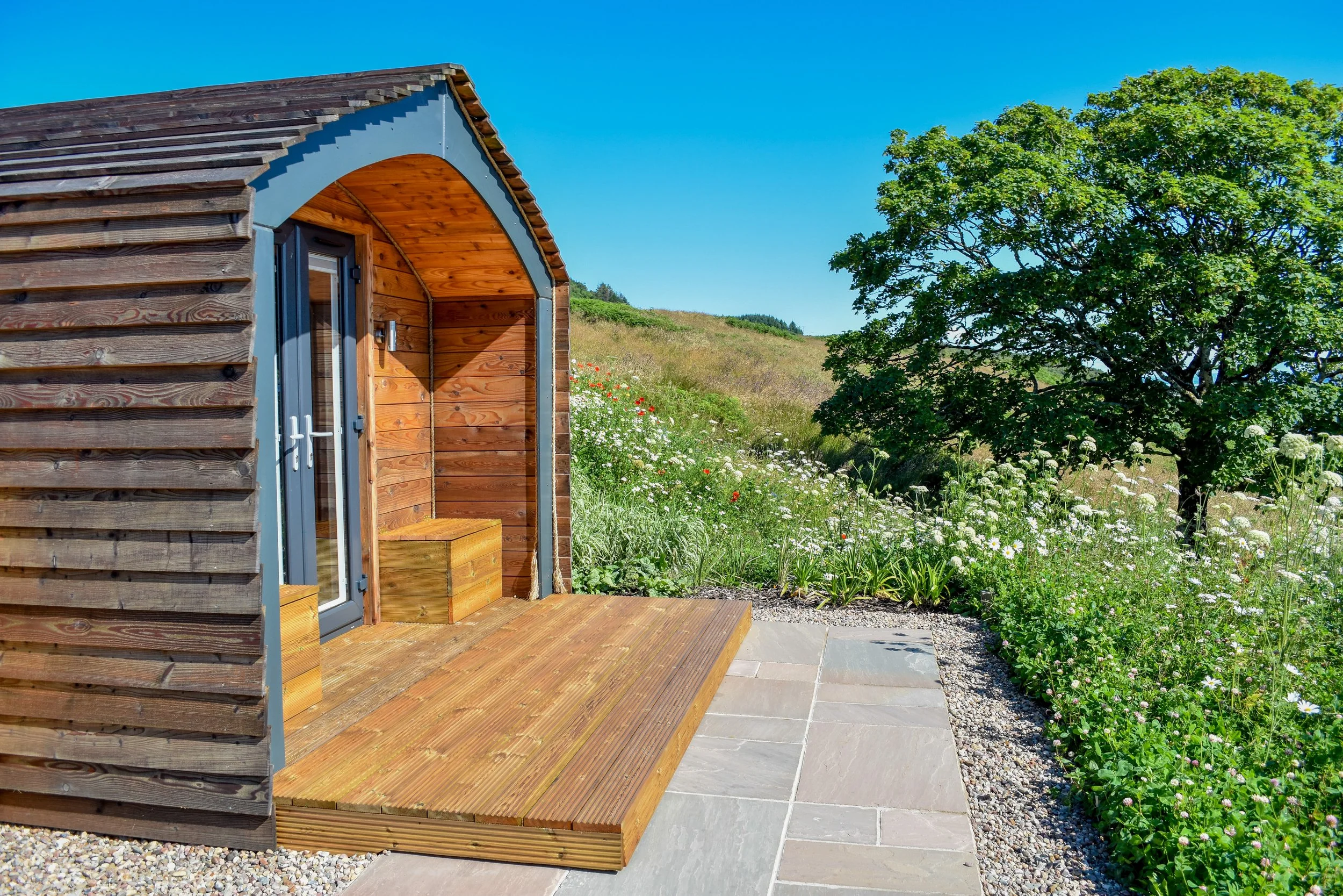 A small wooden cabin with a front porch, set in a scenic natural landscape with grass, wildflowers, and a large green tree under a clear blue sky.