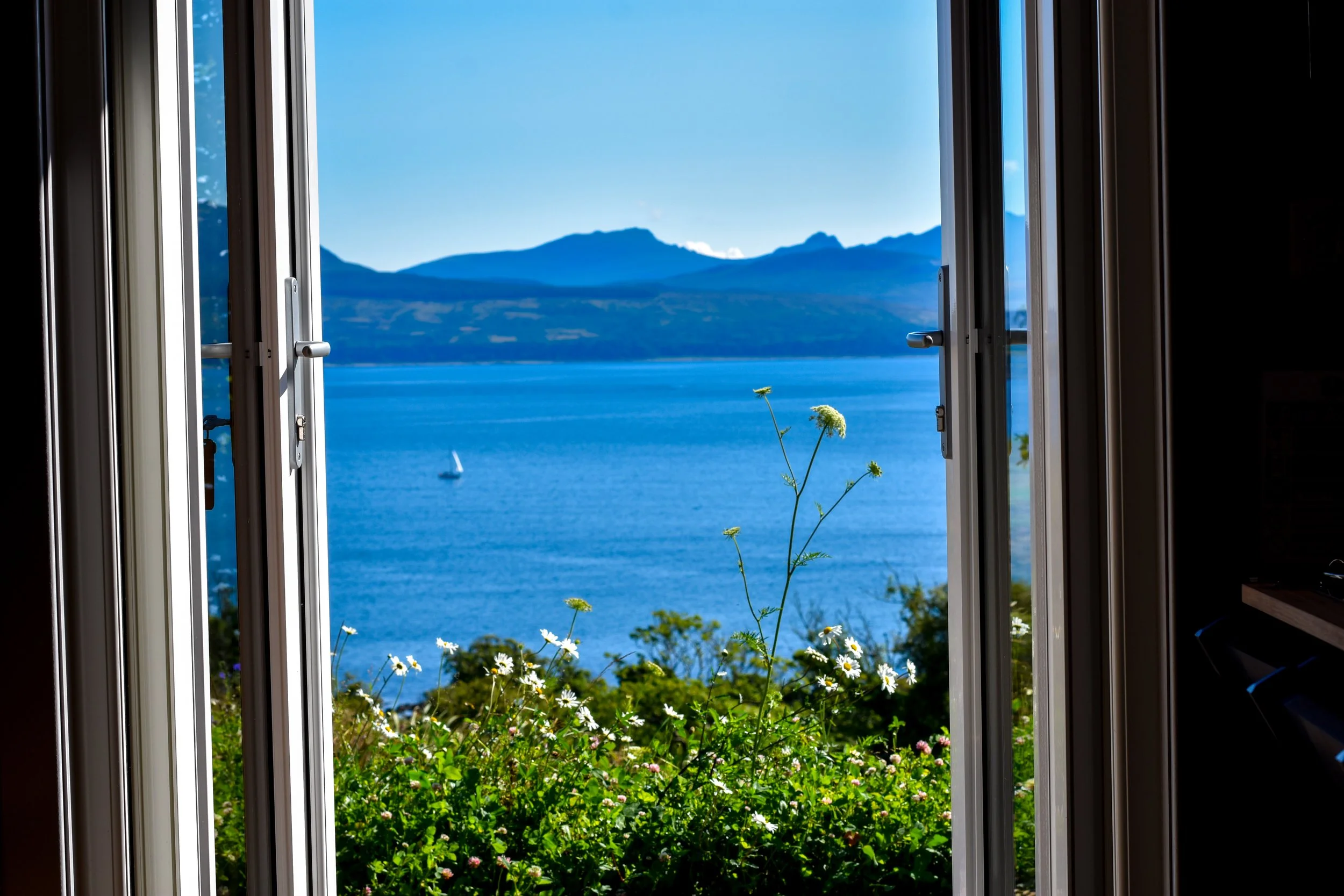 View through an open window showing blue water and mountains in the distance with a sailboat on the lake and flowers in the foreground.