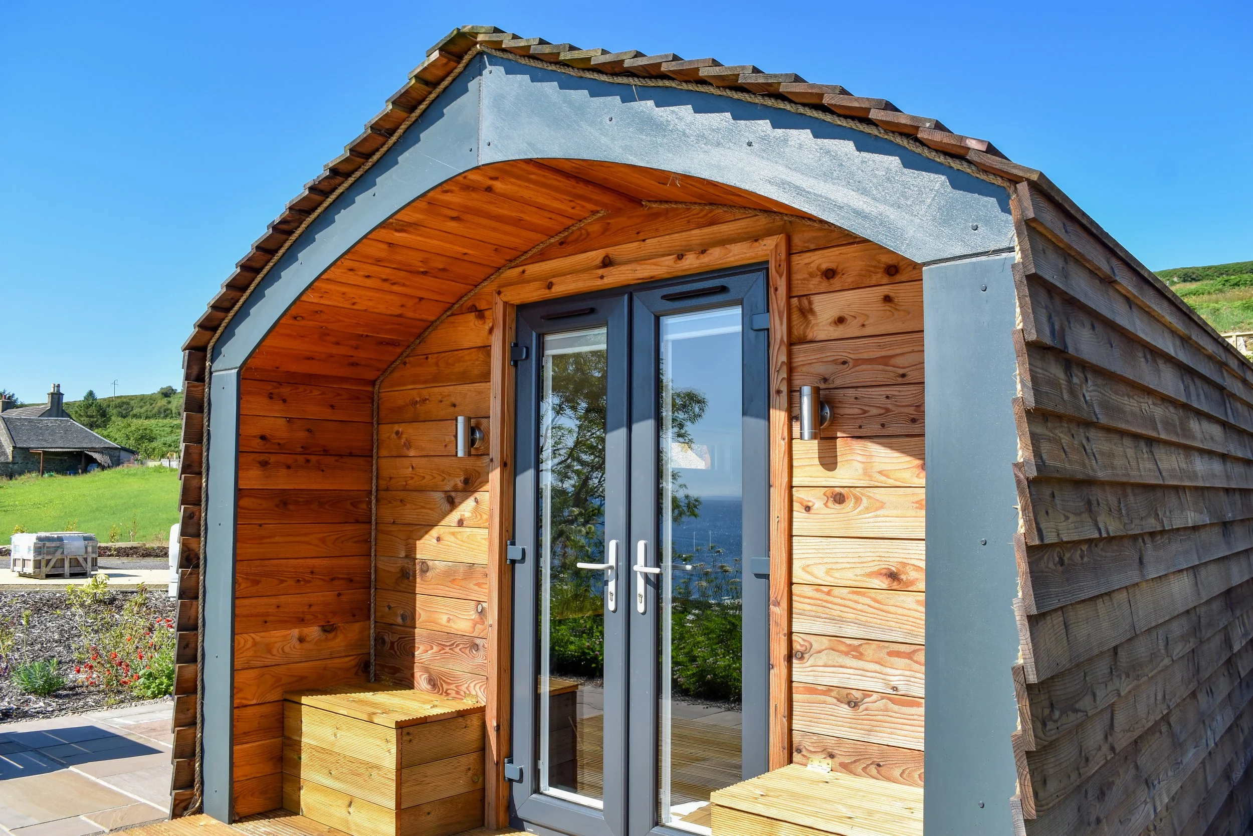 Small wooden garden house or shed with double glass doors, wooden benches, and a curved roof, set in a garden with green grass and hills in the background, under a clear blue sky.