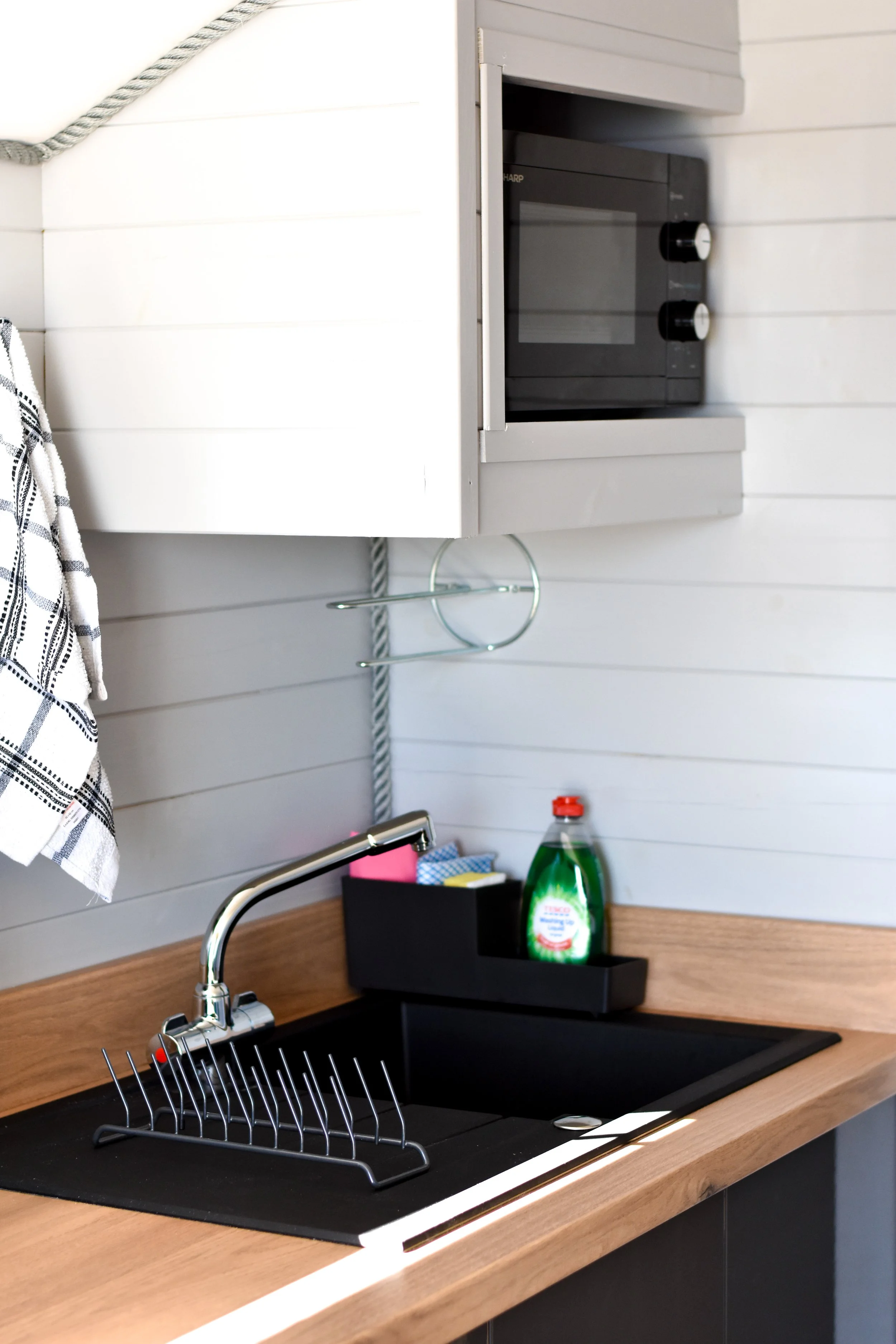 Kitchen sink with dish rack, soap bottle, and dish towel, with microwave on white wall above.