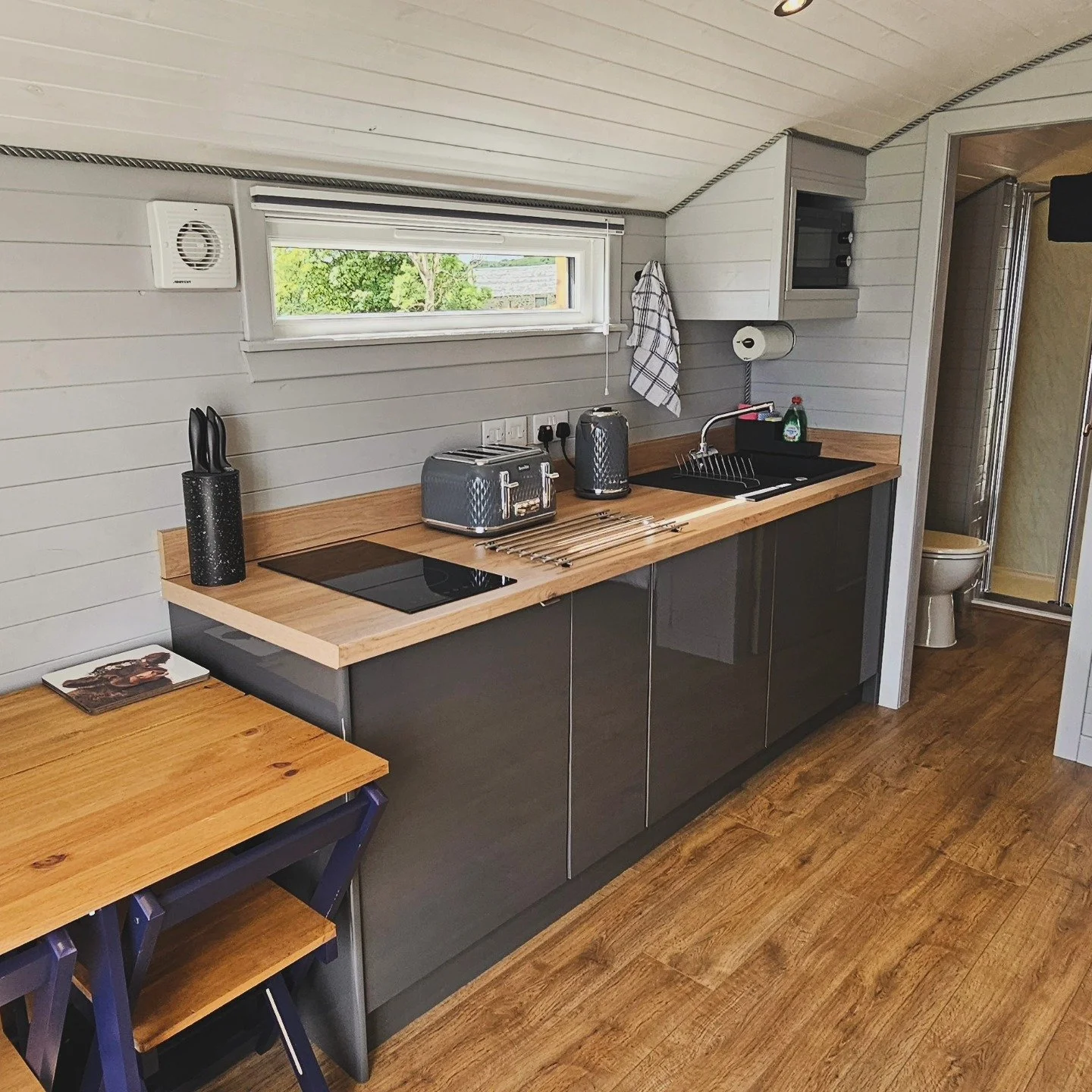Kitchen with black cabinetry, wooden countertop, small window, toaster and kettle on the counter, dish soap near sink, towels, and a small table with a magazine and chairs. Visible bathroom area with toilet and shower.