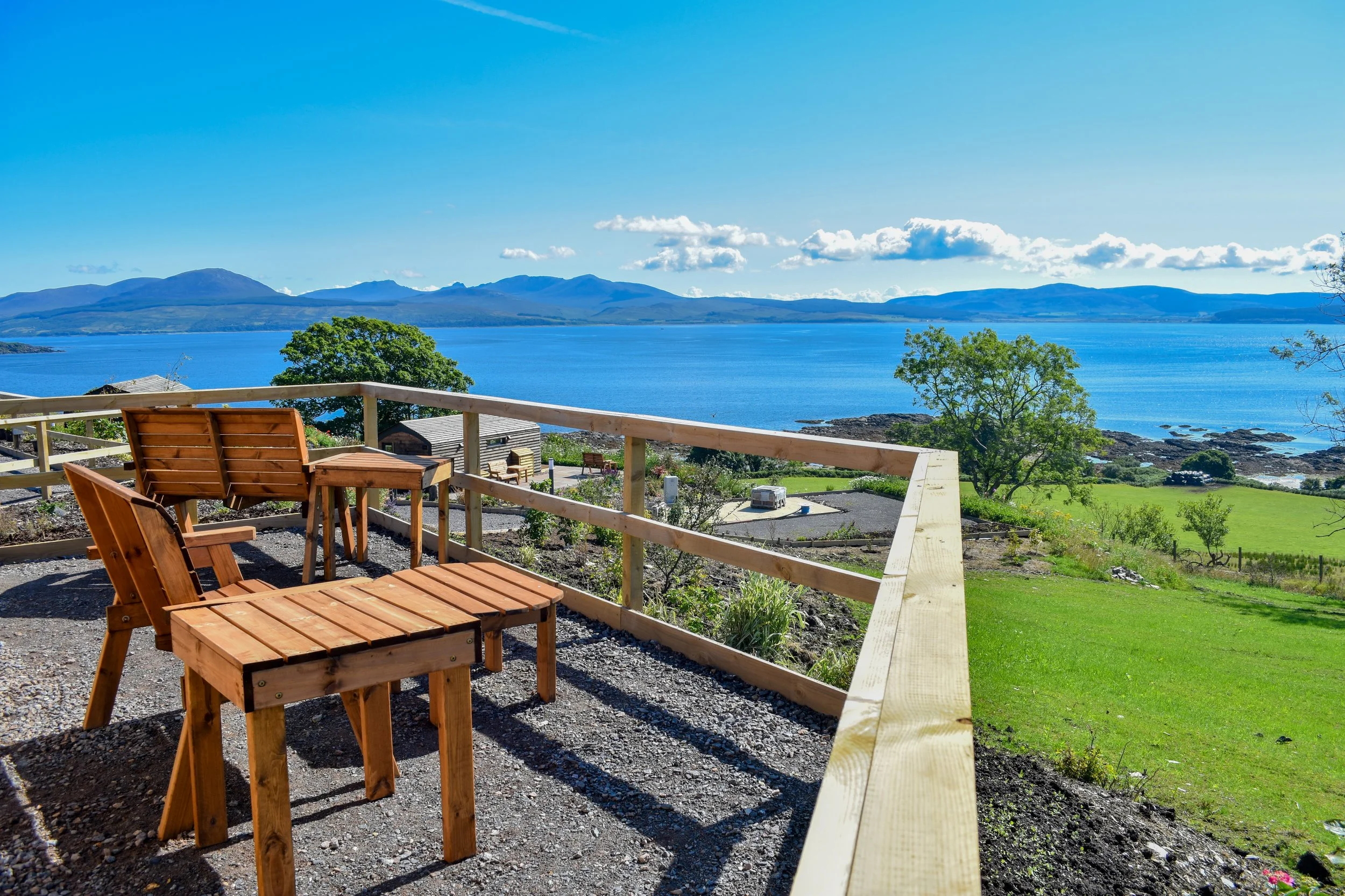 Wooden outdoor seating area on a deck with a lake and mountain view in the background under a blue sky with clouds.