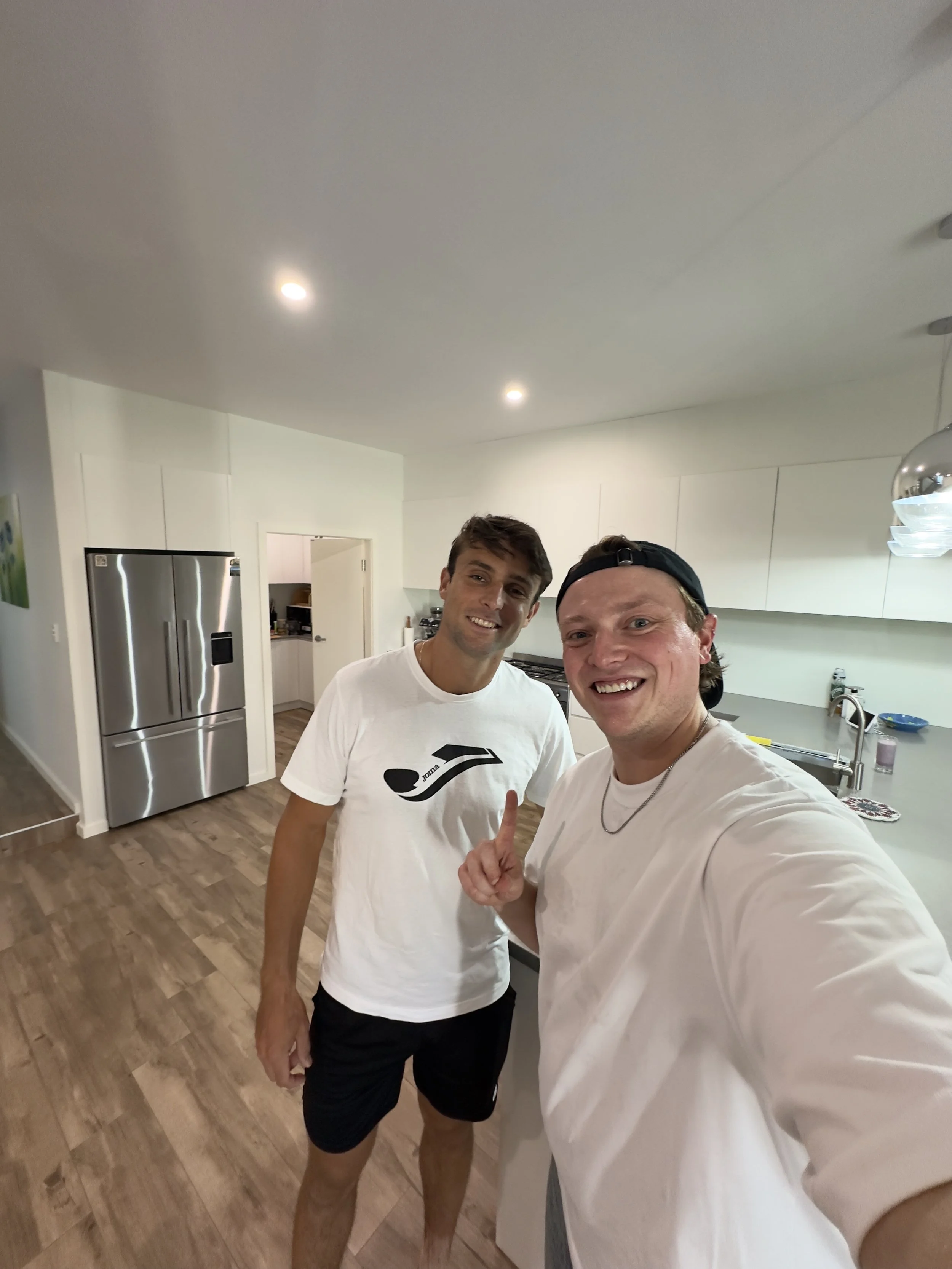 Two young men smiling for a selfie in a modern kitchen, one wearing a white t-shirt with a black logo and the other in a white shirt and backwards cap.