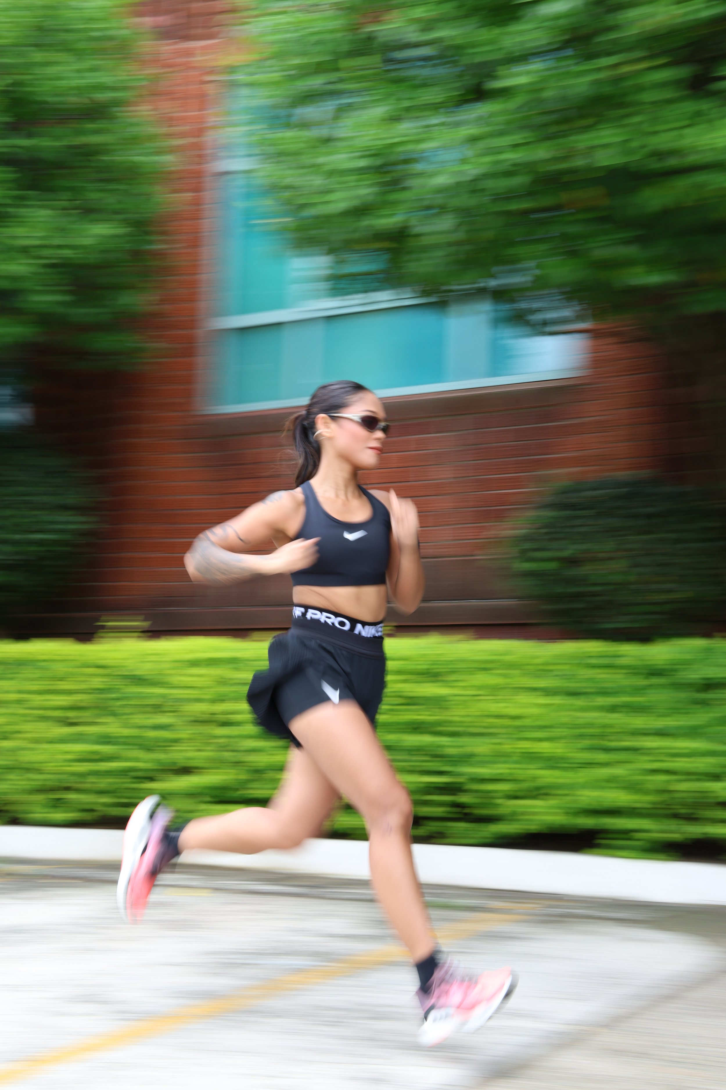 A woman running outdoors in athletic gear with a blurred background of a brick building and green bushes.