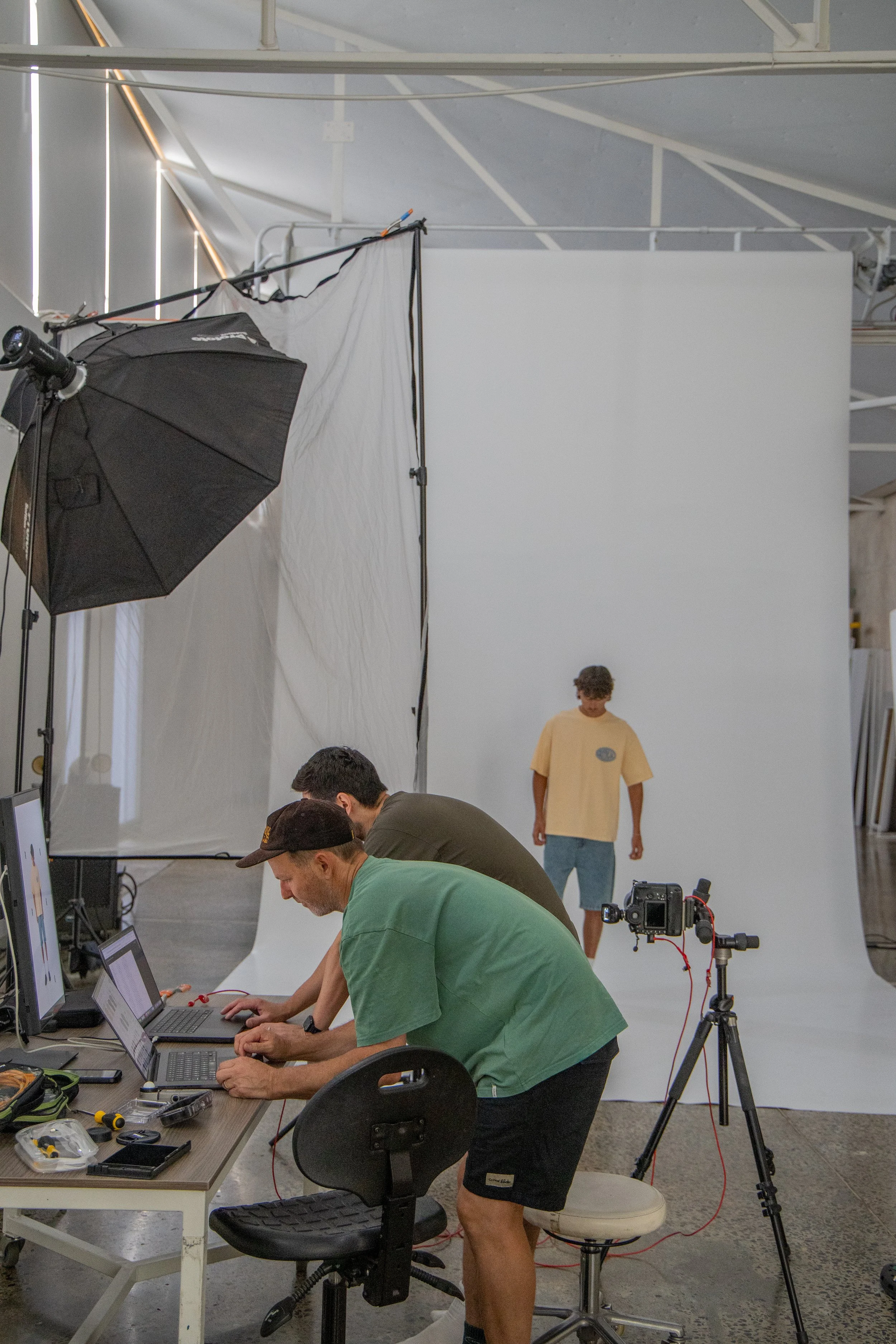 A photo of a photo studio where three people are working with laptops in front of a white backdrop, with one person walking in the background. Photo equipment includes a large umbrella light, multiple tripods, and a camera.
