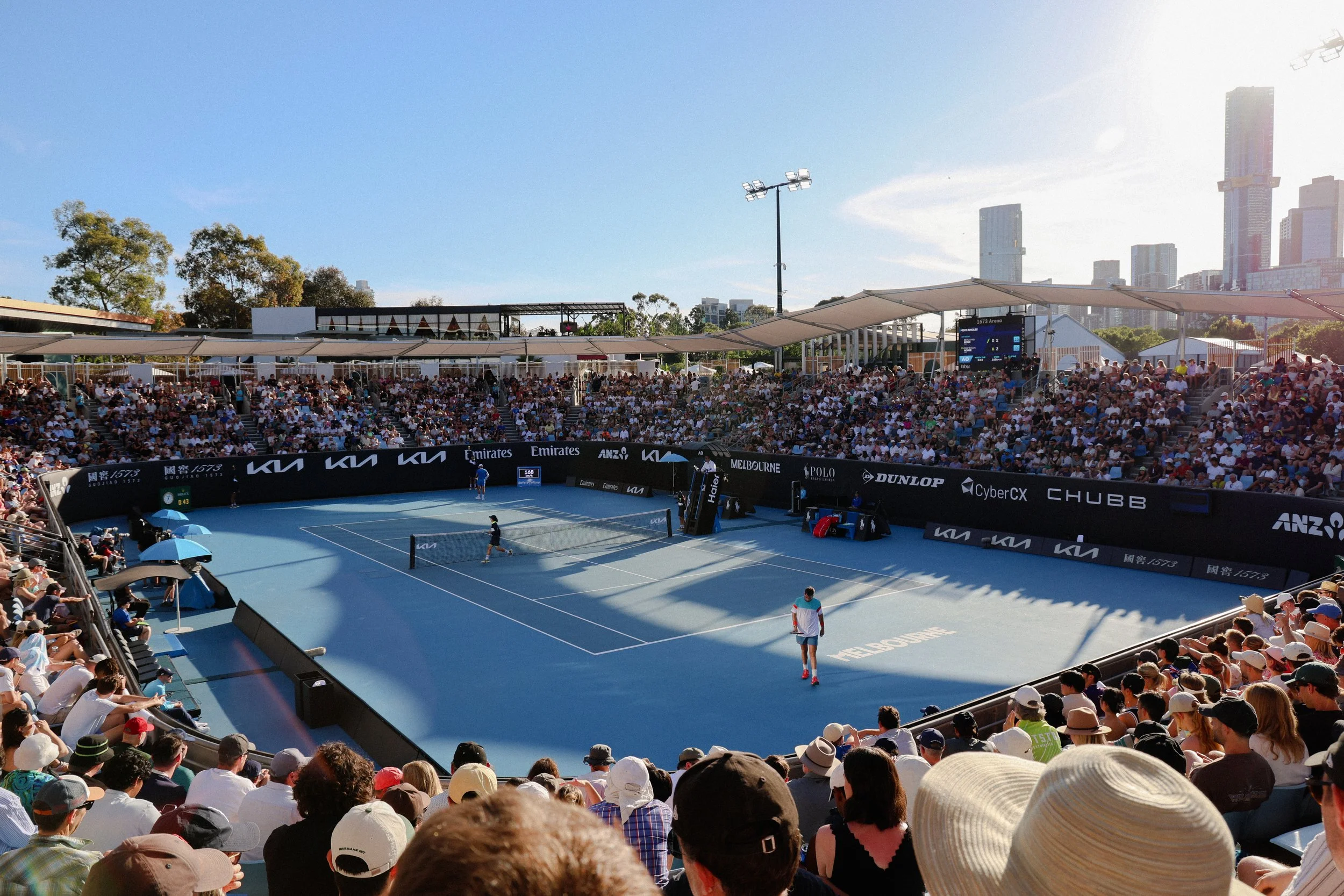 Crowd watching a tennis match at an outdoor stadium in Melbourne on a sunny day.