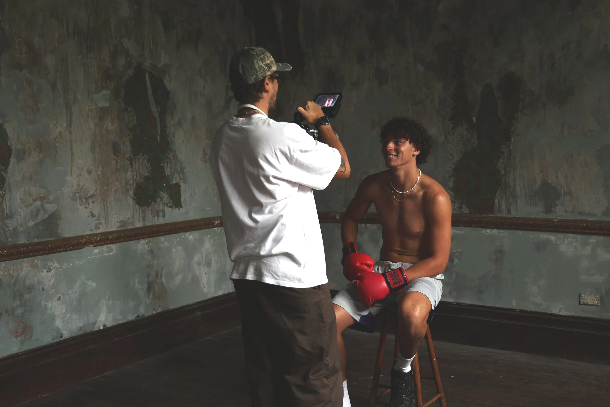 A man sitting on a stool with red boxing gloves, shirtless, wearing shorts, being photographed by a person holding a camera in an old, dimly-lit room with peeling paint.
