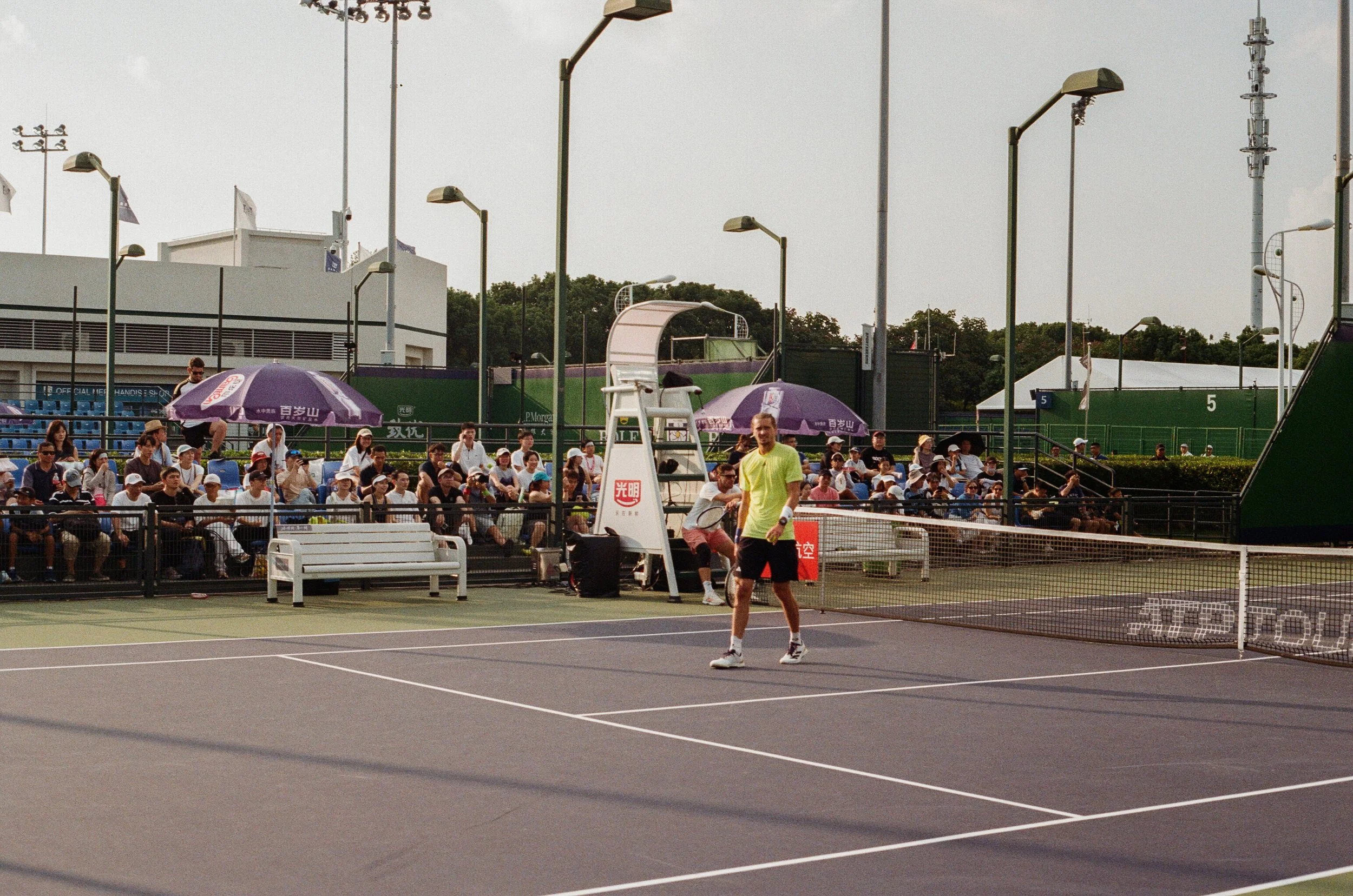 A man in a yellow shirt and black shorts on a tennis court, with a crowd of spectators sitting on bleachers in the background under purple umbrellas, and a sunny sky overhead.