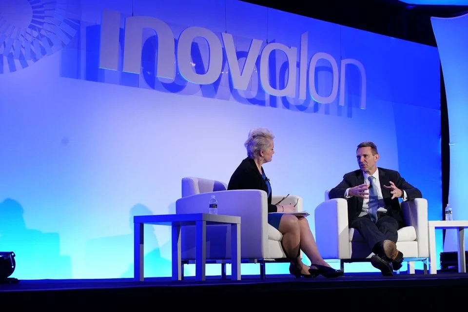 Two people sitting on stage in conversation at a conference, with a large 'Inovation' banner in the background, engaged in discussion, with water bottles on small tables beside them.