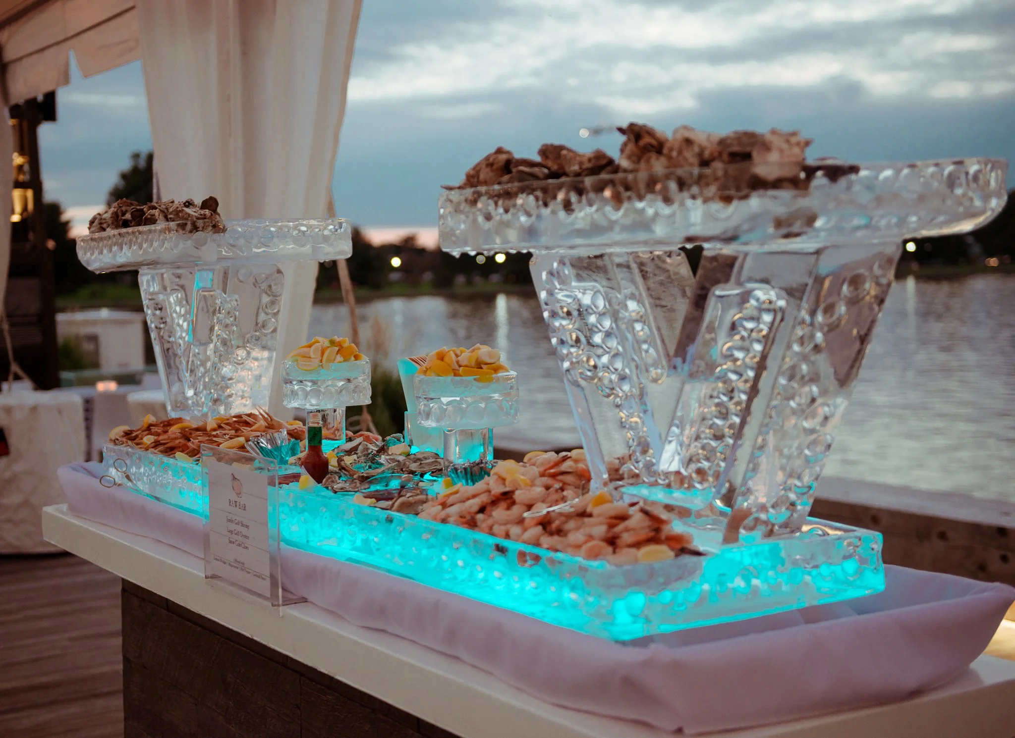 Ice sculpture with seafood and snacks, illuminated with blue light, set near a water body at sunset.