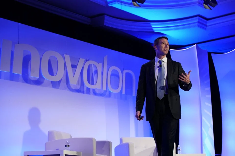 A man in a black suit giving a presentation on stage with a large blue 'Innovación' sign and blue lighting.