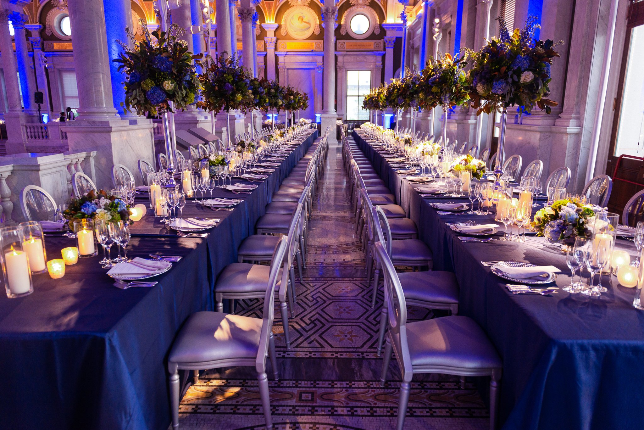 Elegant banquet hall with long tables decorated with floral centerpieces, candles, and glassware, set for a formal event or wedding reception.