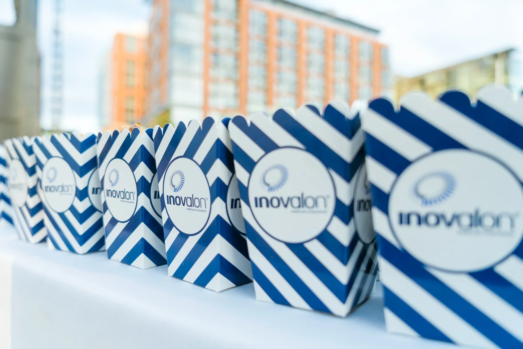 Rows of blue and white patterned gift bags with the 'Inovalon' logo printed on them, set outdoors with a blurred cityscape background.