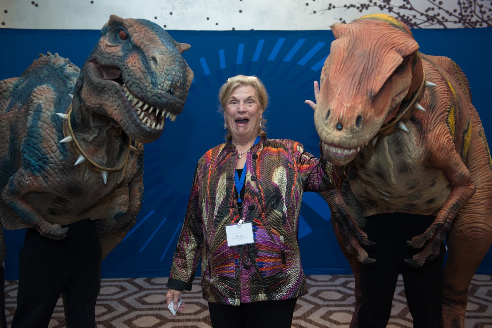 A woman standing between two people in dinosaur costumes, smiling and holding up her right hand with a mischievous expression.
