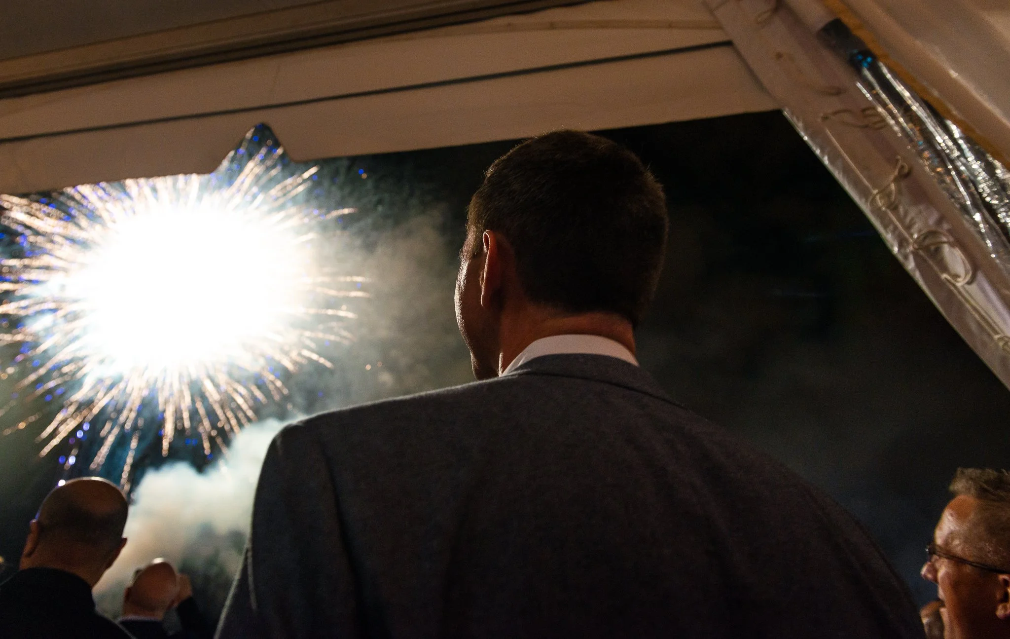 A man in a suit watching fireworks at night from inside a building with metallic and wooden interior.