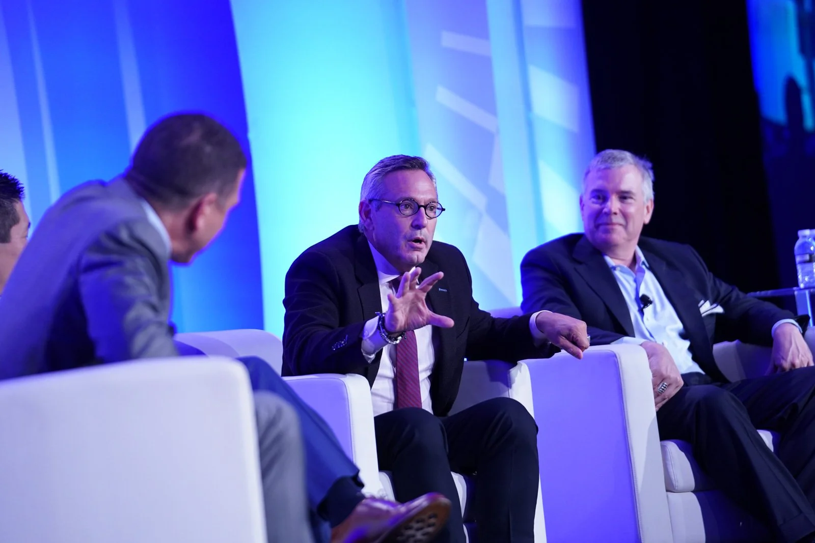 Four men in suits sitting on a panel at a conference; one speaking and gesturing with his hand, others listening.