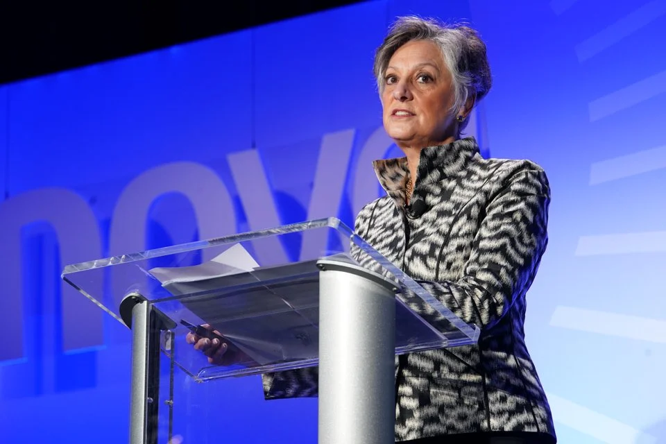 A woman with gray hair wearing a black and white animal print blazer speaking at a podium during a conference or event.