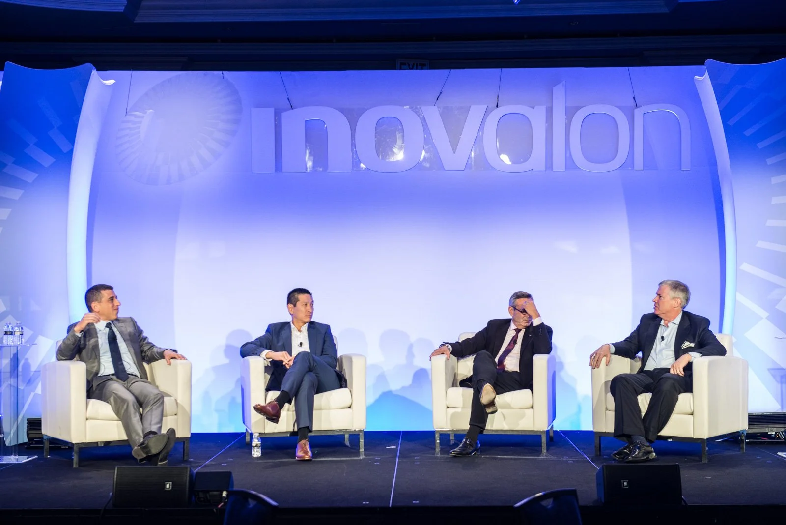 Four men in suits sitting on white chairs on a stage during a panel discussion at an event with a blue backdrop and large 'innovation' sign.