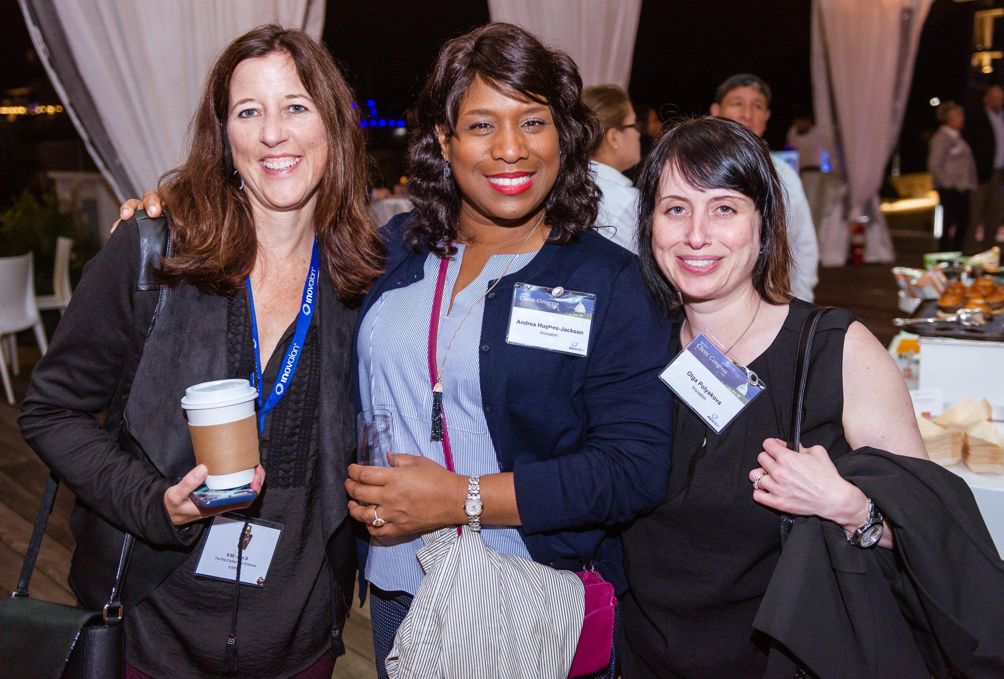 Three women at a social event, smiling and posing for a photo, in an evening setting with people and tables in the background.