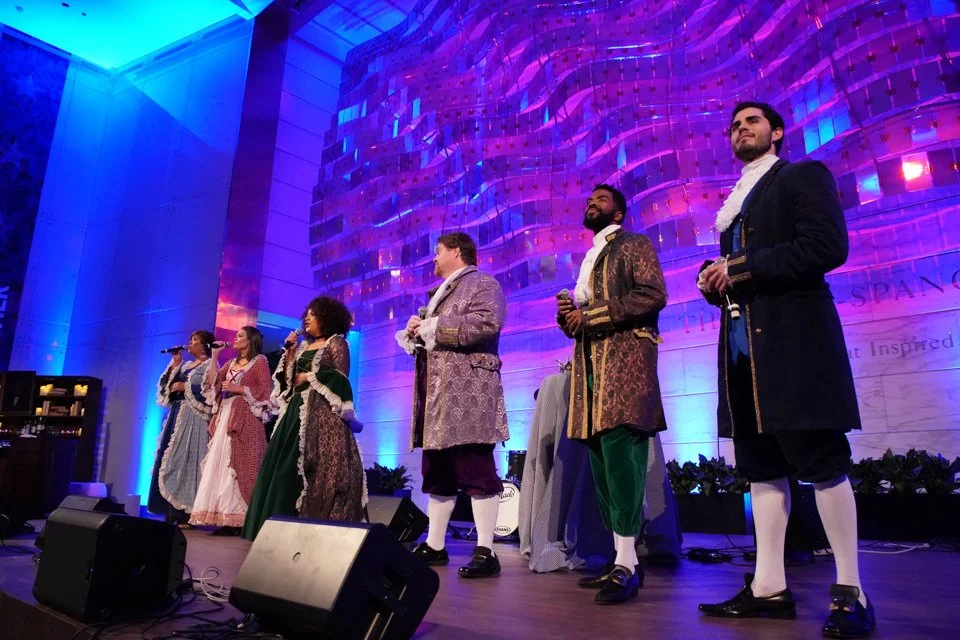 Group of six performers dressed in historical costumes singing on stage with a modern, colorful architectural backdrop.