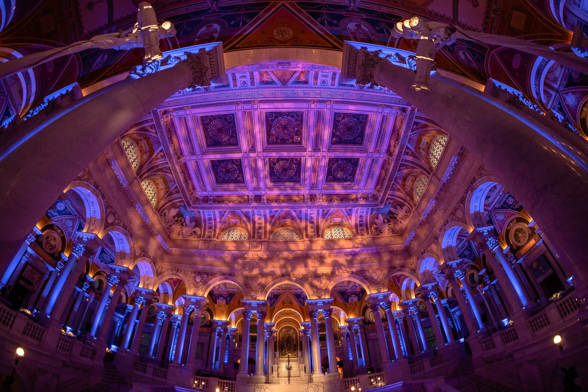 Interior view of a grand hall with ornate architecture, illuminated with purple, blue, and warm lighting, showcasing tall columns, decorated arches, detailed ceiling, and staircase.