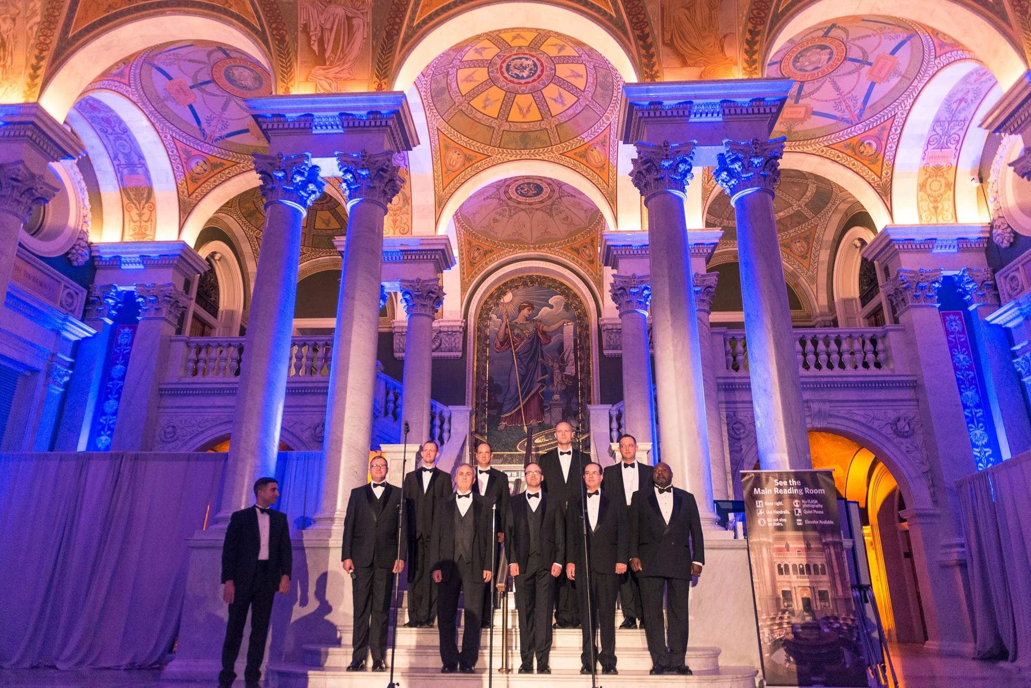An all-male choir dressed in black tuxedos with bow ties performing on the stairs of a grand, ornate hall with tall marble columns, colorful curved ceiling, and a large religious-themed painting in the background.