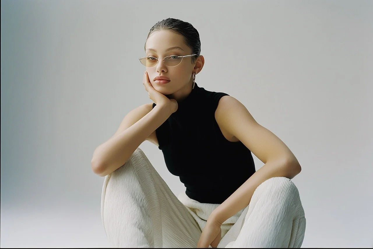 A young woman sitting cross-legged against a neutral background, wearing glasses, a sleeveless black top, and beige pants, with her hand resting on her face.