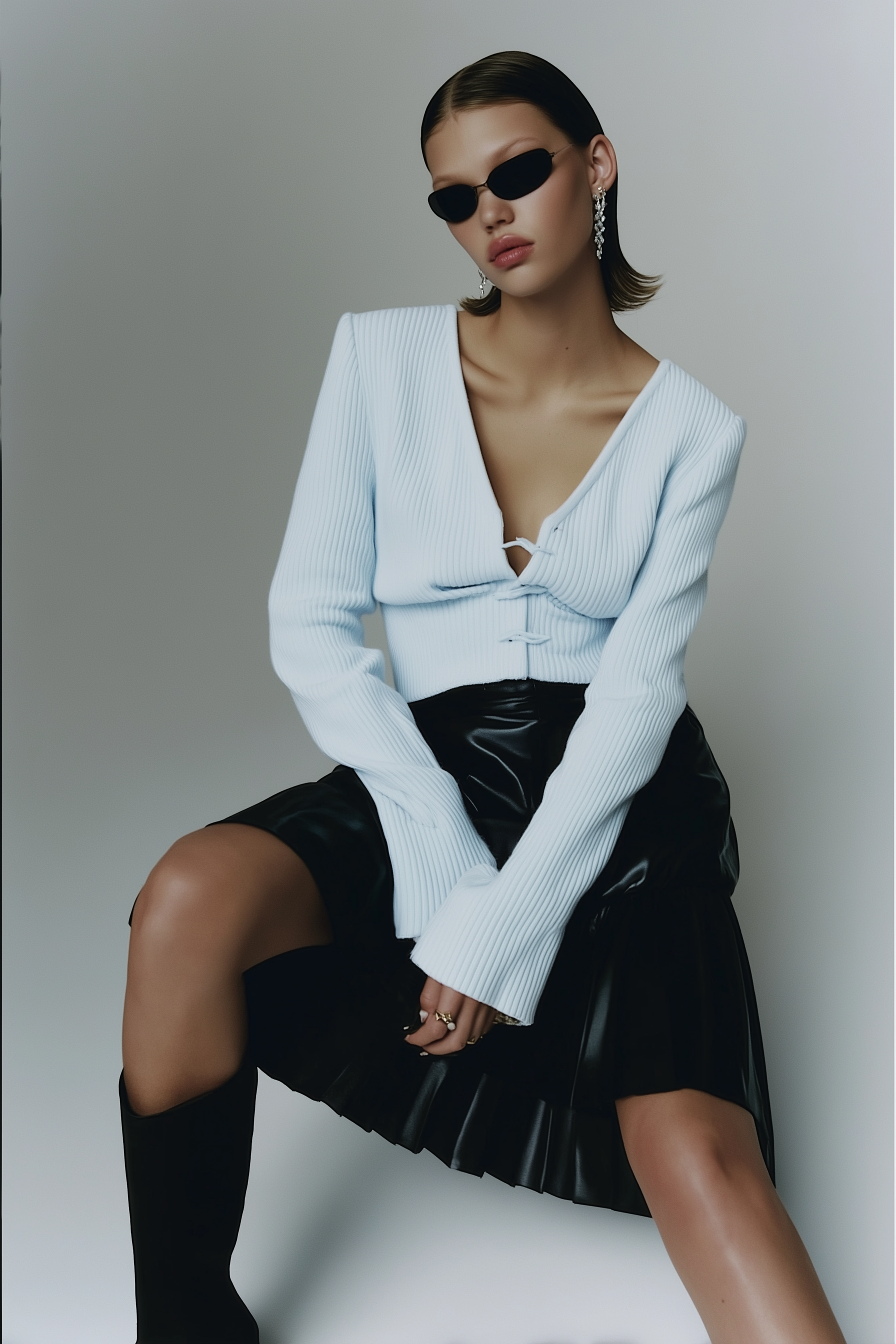 A young woman with slicked-back hair, wearing black sunglasses, a white long-sleeve ribbed top, a black leather pleated skirt, and black knee-high boots, sitting against a neutral background.