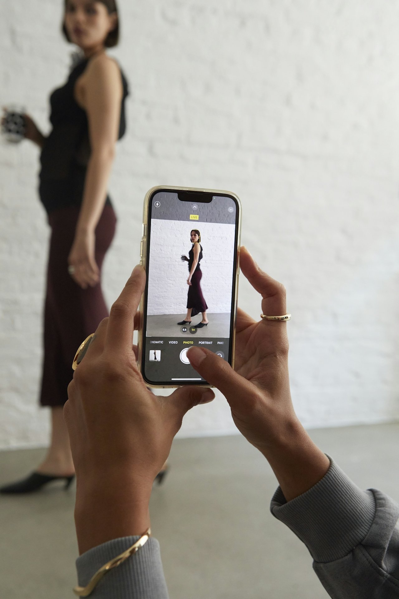 Person holding a smartphone taking a photo of a woman standing against a white brick wall. The woman's image appears on the phone screen.