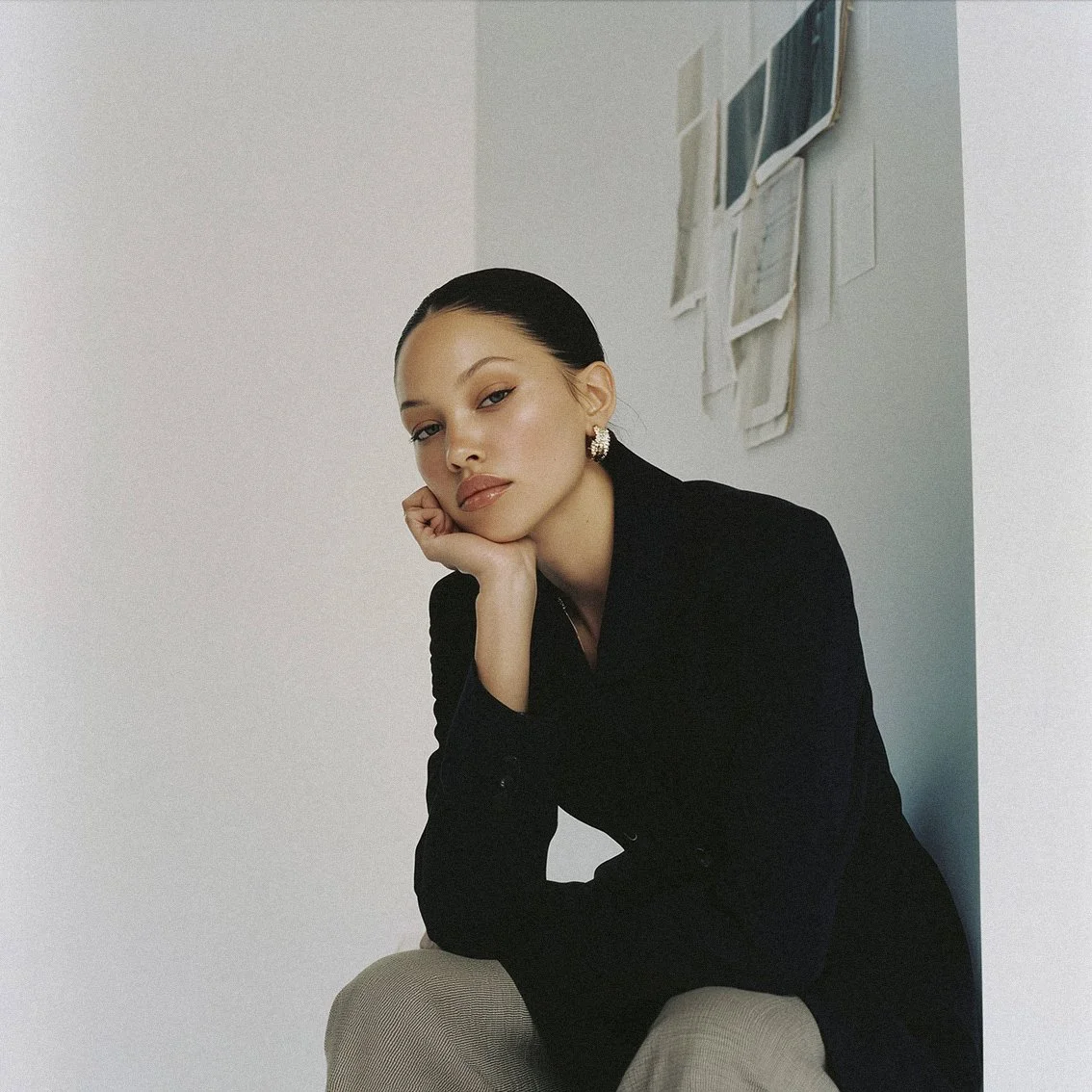A woman with dark hair pulled back, wearing a black blazer and earrings, sitting against a white wall with some papers pinned or taped on it.
