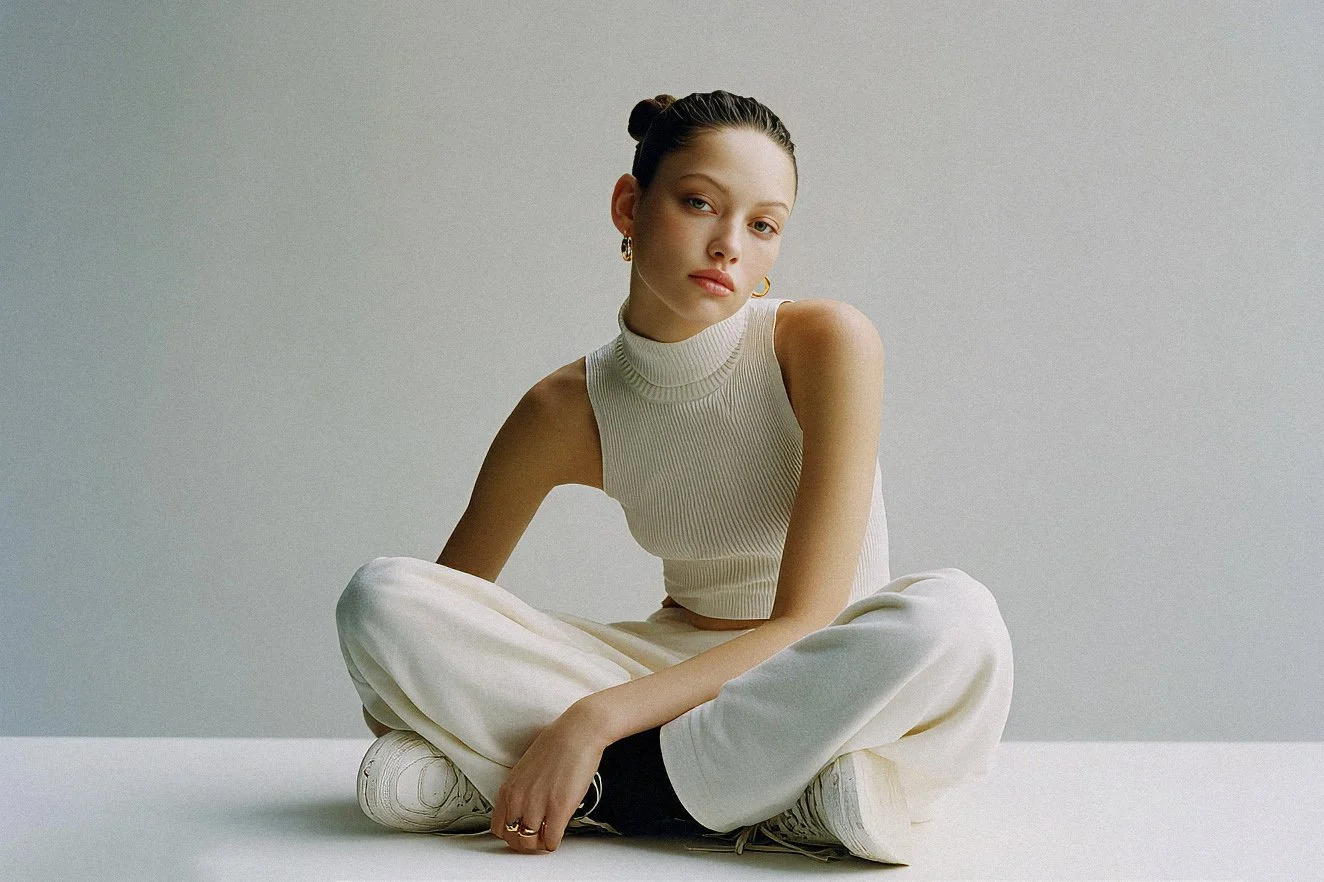 Young woman sitting cross-legged on the floor against a plain, light-colored background. She has dark hair styled in a bun and is wearing a sleeveless, high-neck, ribbed top, cream-colored pants, and white sneakers. She has gold hoop earrings and a subtle, natural makeup look.